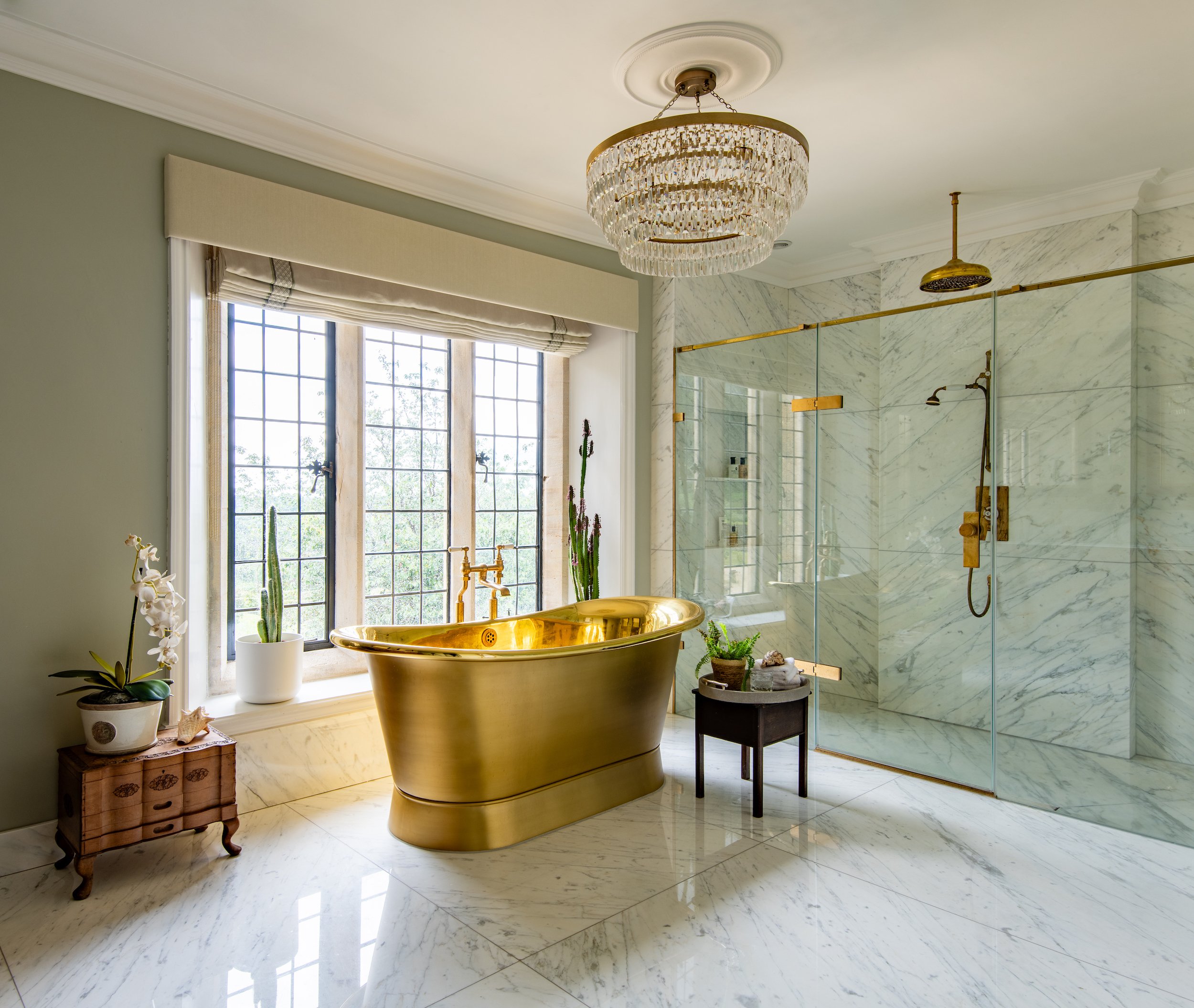 Master bathroom designed by MIDA Interiors for a large Cotswolds country estate, featuring a statement brass freestanding bath, bookmatched marble flooring and wall cladding, a generous walk-in shower, bespoke brass fittings and a crystal chandelier.
