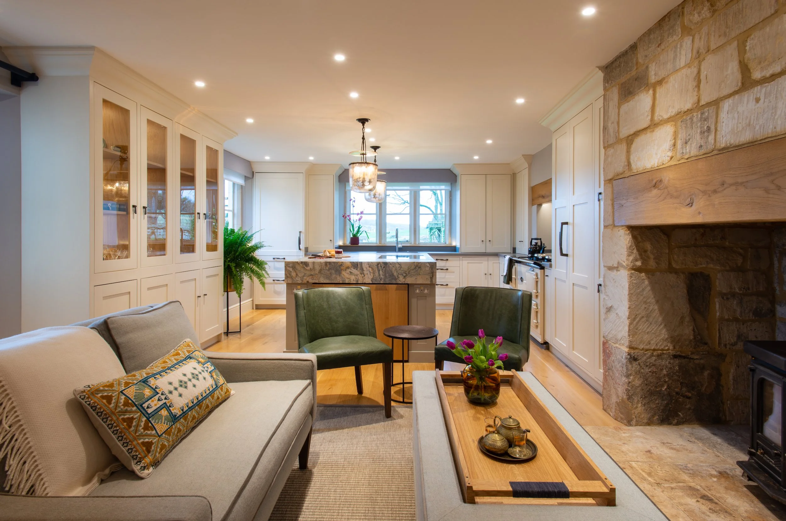 A wide view of the replanned kitchen and adjoining fireside seating within a Grade II listed Cotswold farmhouse. Bespoke painted cabinetry, new oak flooring and a quartz waterfall island create a refined architectural framework, while tailored seatin