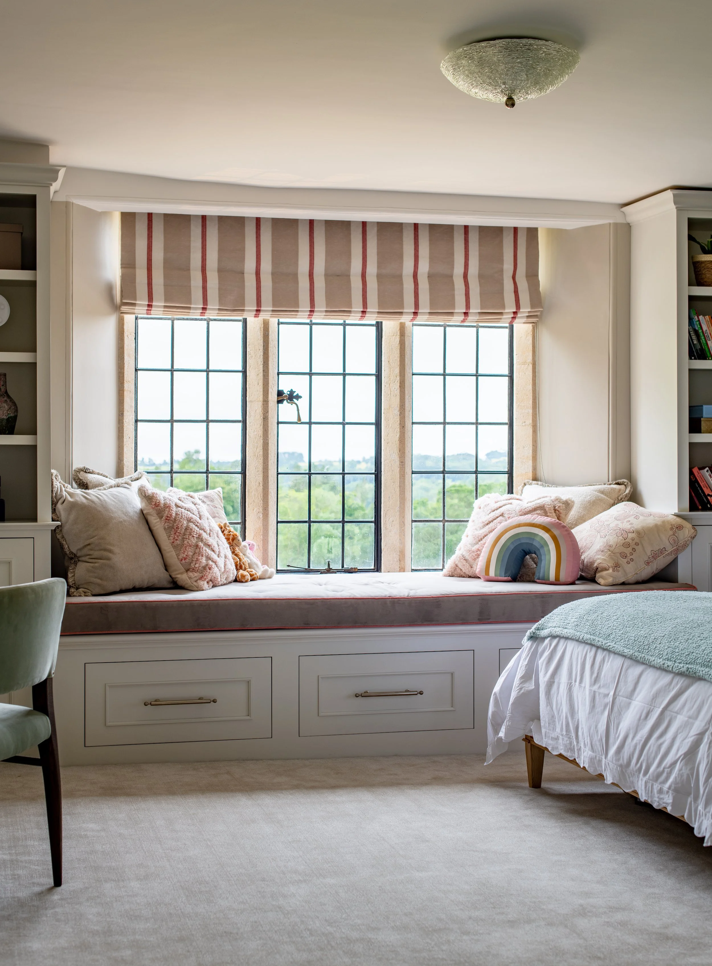 Children’s bedroom window seat designed by MIDA Interiors for a large Cotswolds country estate, featuring bespoke under-seat storage, integrated shelving, tailored soft furnishings and expansive views across the grounds.