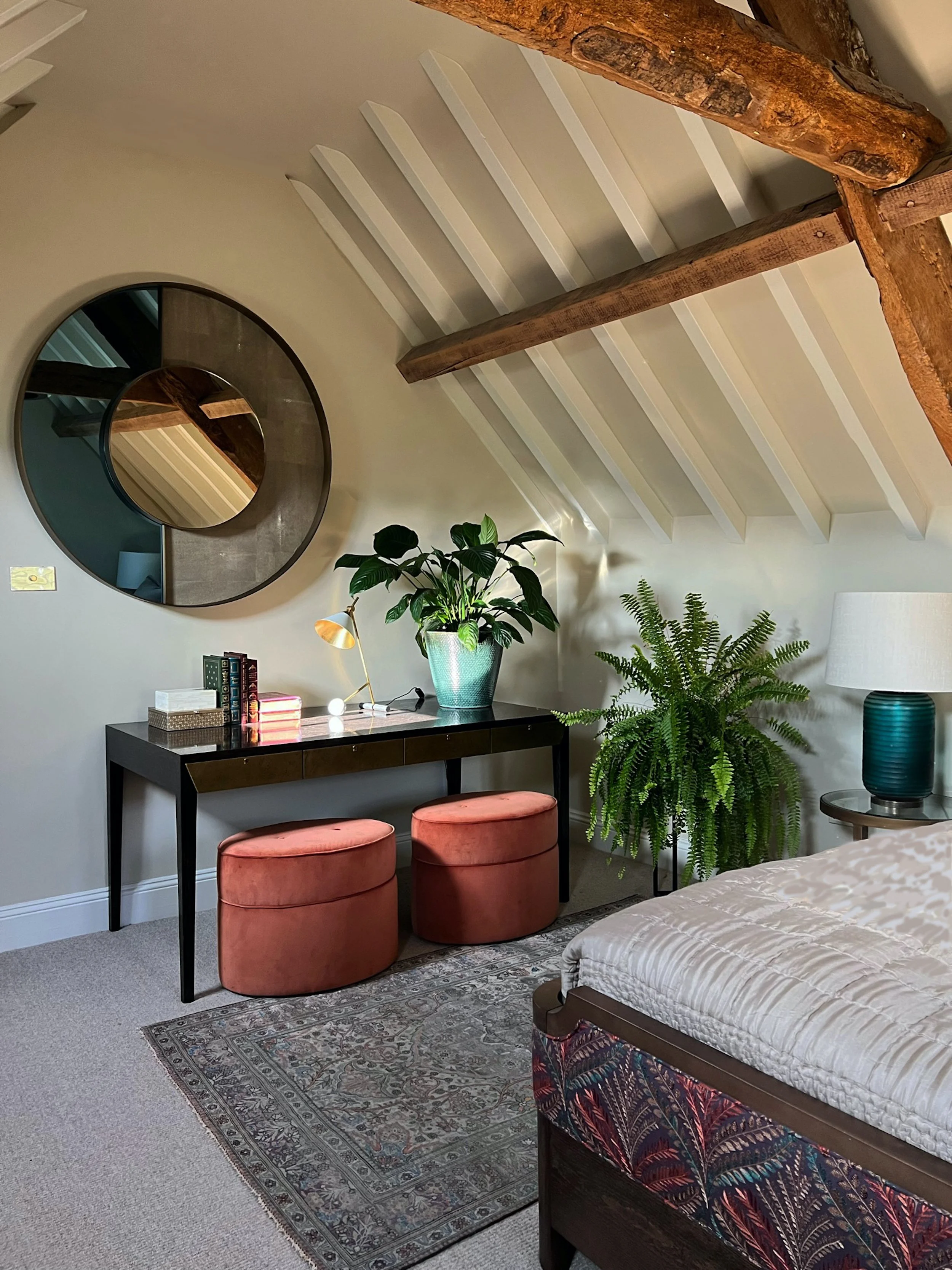 Guest bedroom dressing area beneath exposed beams, with a dark dressing table, round mirror, coral upholstered stools and indoor planting.