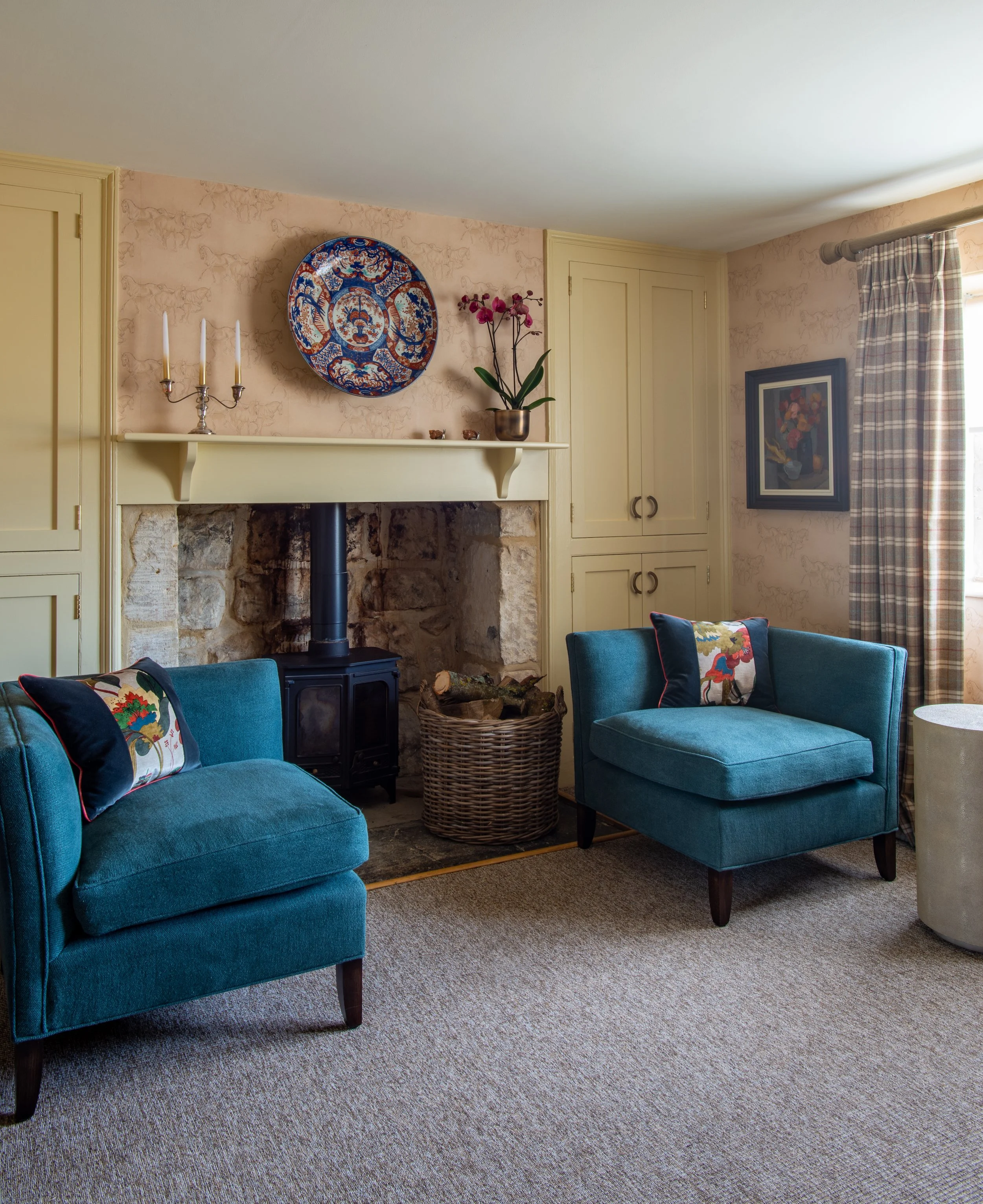 The flexible seating area within the farmhouse study features two bespoke teal armchairs arranged around a stone fireplace and wood-burning stove. An antique Imari porcelain charger creates a distinctive focal point above the mantel, complemented by 