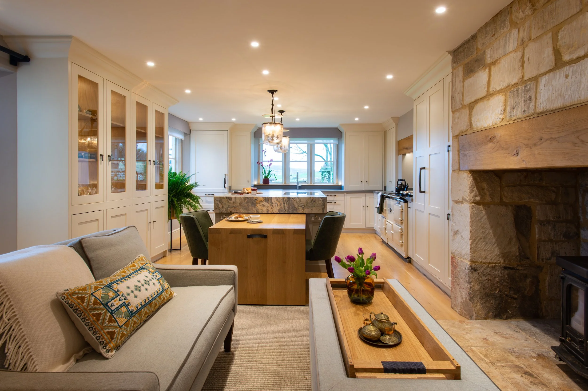 Bespoke kitchen and living space designed by MIDA Interiors for a Cotswolds country house, featuring an oak dining table that slides beneath the island to create flexible seating for intimate meals or larger gatherings.
