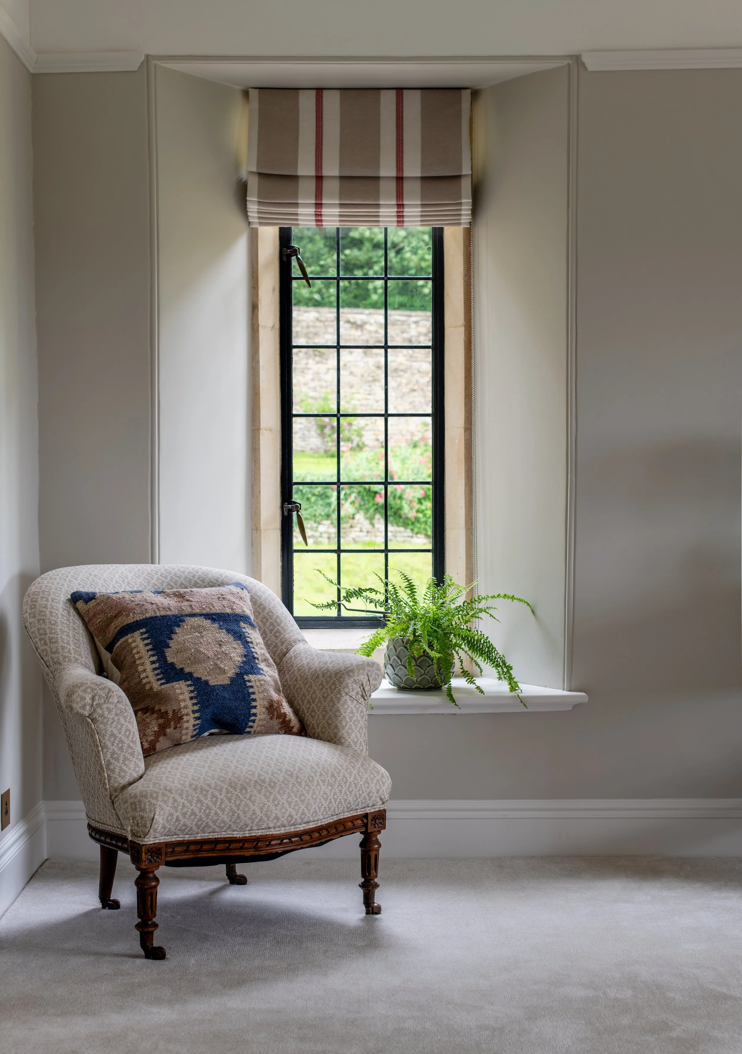 Children’s bedroom reading corner designed by MIDA Interiors for a large Cotswolds country estate, featuring an antique upholstered chair, tailored Roman blind and a quiet window-side setting overlooking the garden.