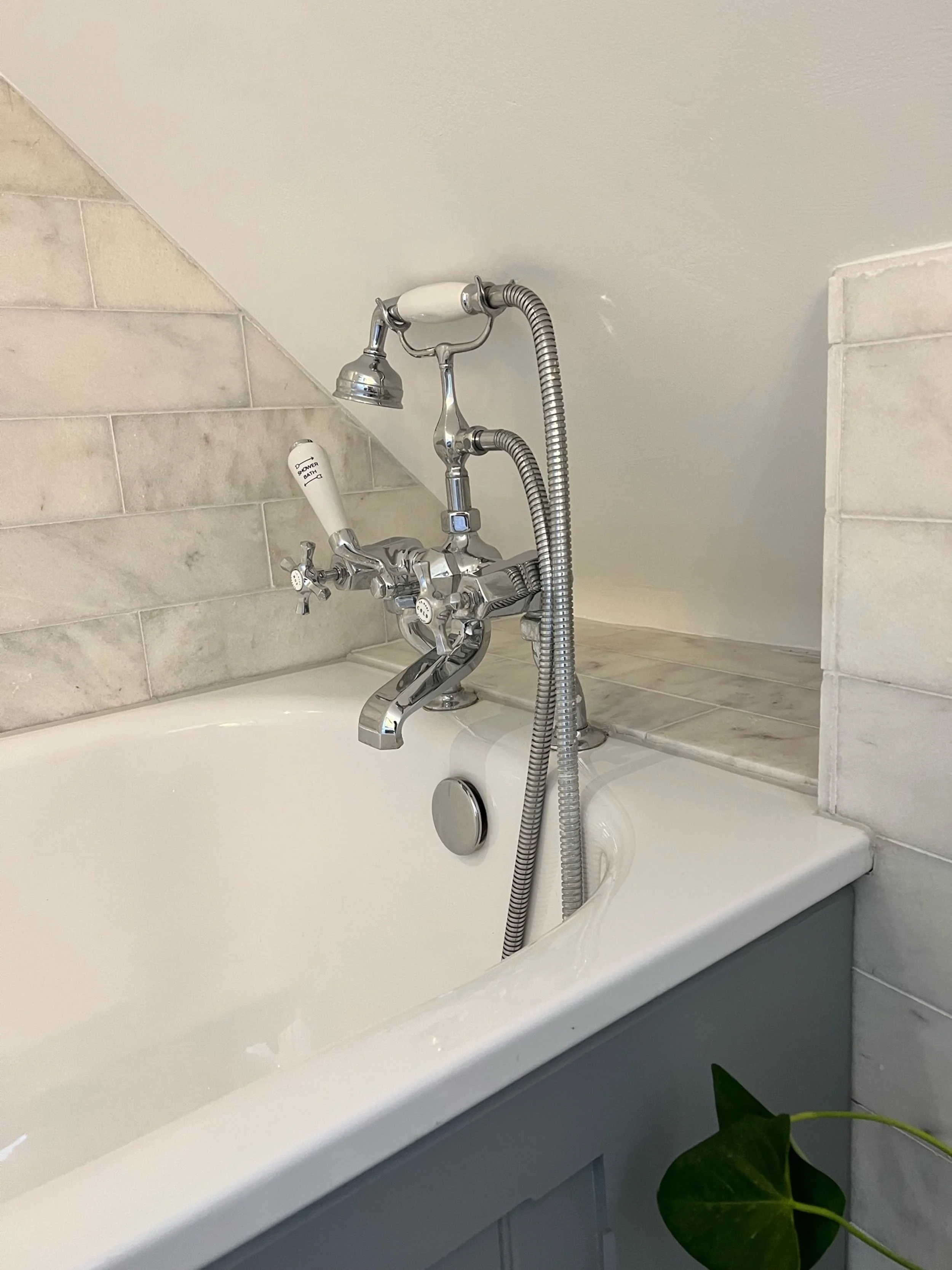 Traditional chrome bath fittings were selected to complement the understated character of the guest bathroom. The bath is set beneath the sloping ceiling and framed by softly veined marble tiles, allowing the compact space to feel coherent and approp