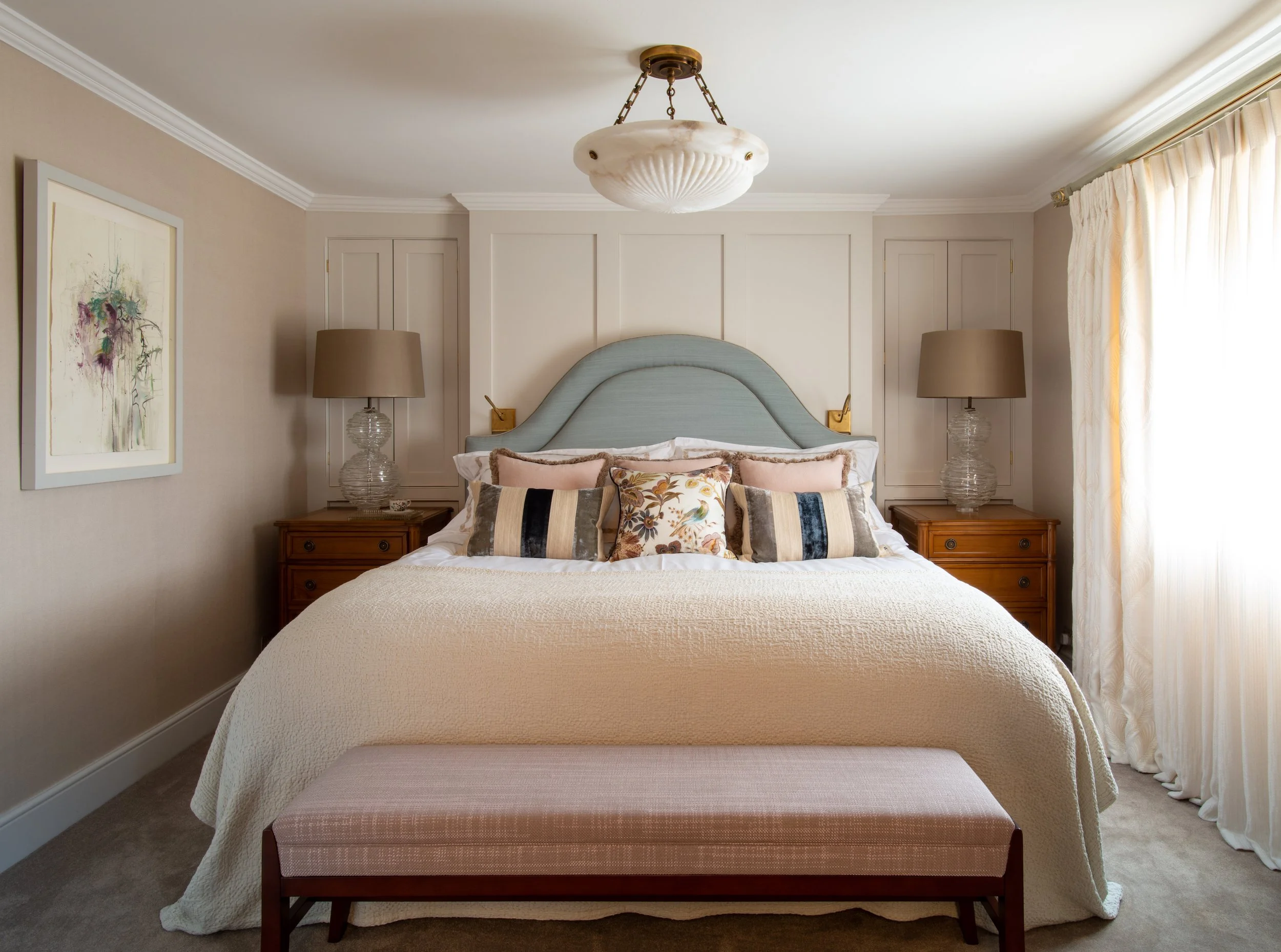The principal bedroom within the reconfigured Cotswold farmhouse suite is centred on a tailored pale blue upholstered headboard set against restrained panelled joinery. Symmetrical bedside chests and glass lamps bring order to the composition, while 