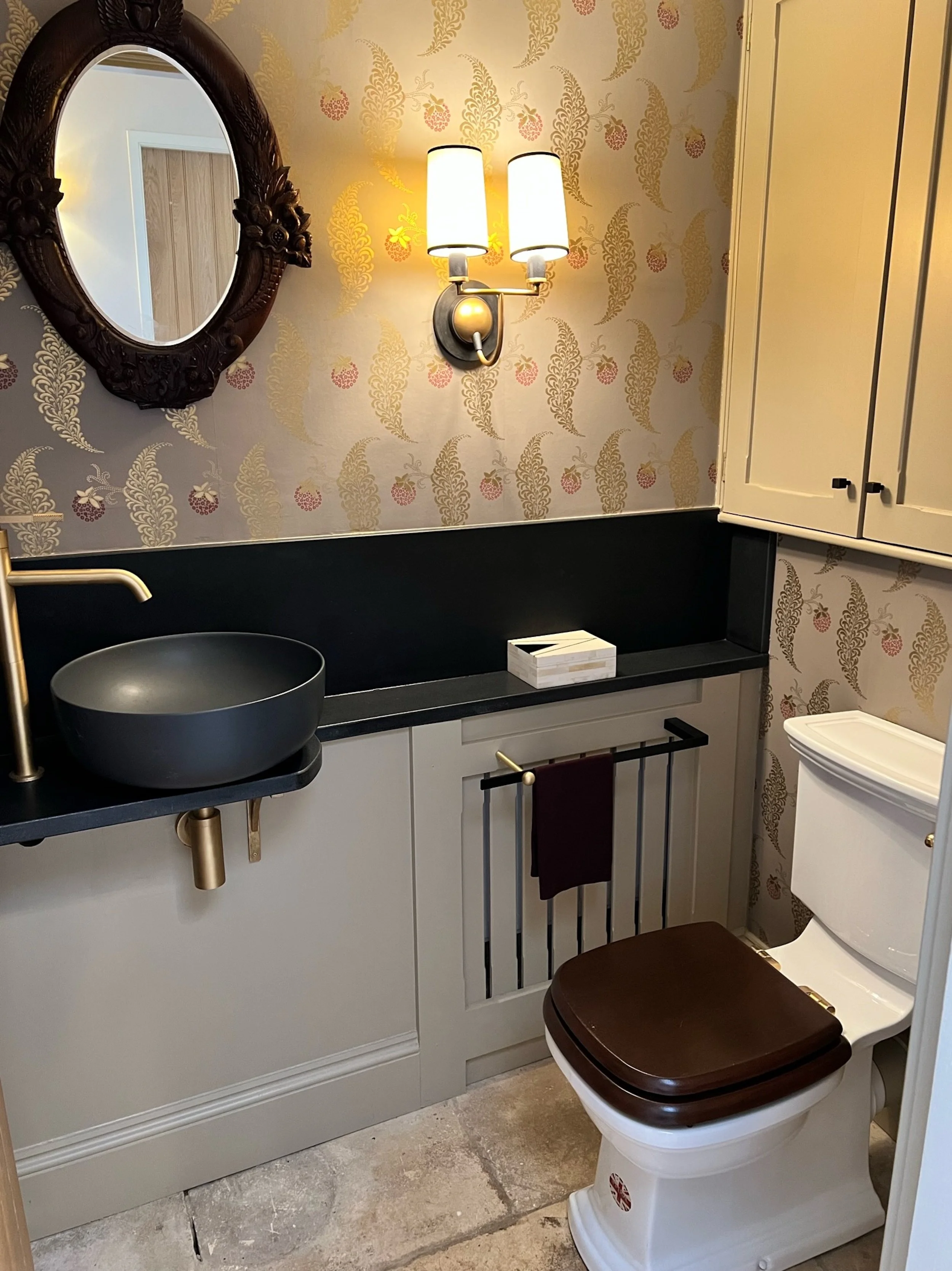 Compact cloakroom within a Cotswold country house, combining patterned botanical wallcovering with bespoke painted joinery and a restrained dark stone surface. A sculptural countertop basin, aged-brass fittings, an antique carved mirror and softly di