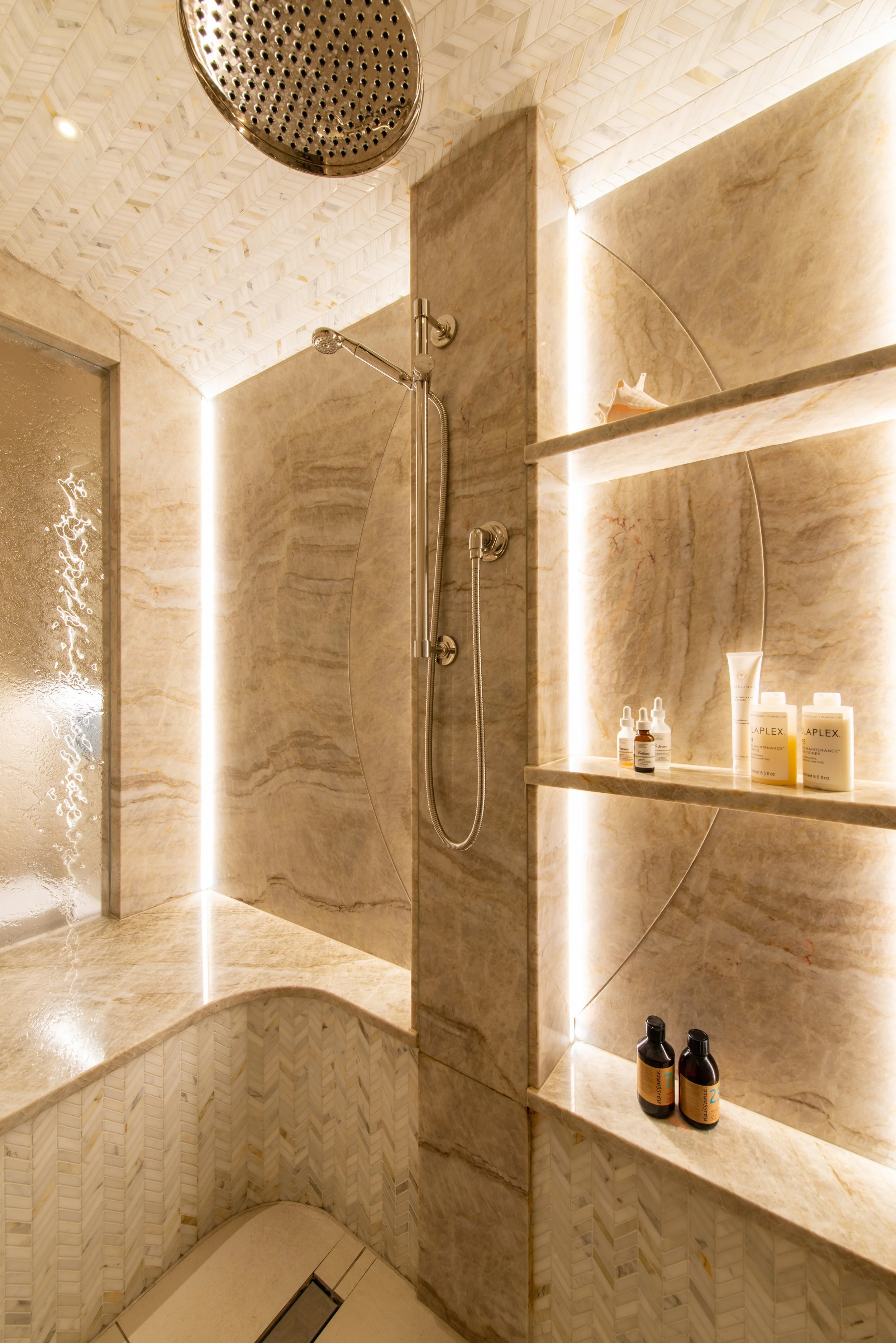 The bespoke steam shower is lined with softly veined stone and a pale herringbone mosaic, combining generous proportions with carefully resolved detailing. A rainfall showerhead, handheld spray, integrated bench and illuminated shelving support every