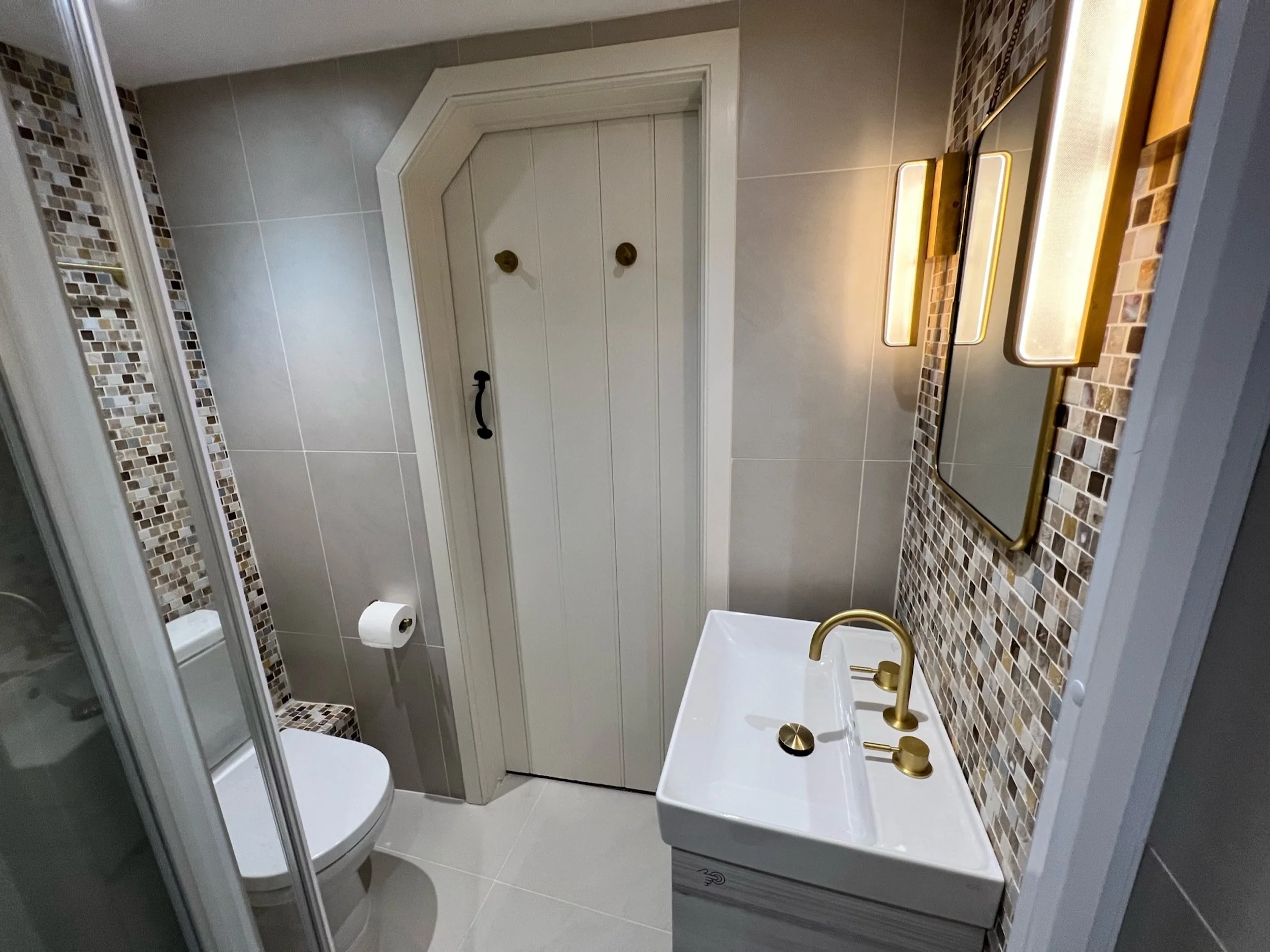 Careful spatial planning allows the principal guest shower room to accommodate a walk-in shower, vanity and WC within its compact proportions. Large format grey tiles create a calm architectural backdrop, while mosaic detailing, warm metal finishes a
