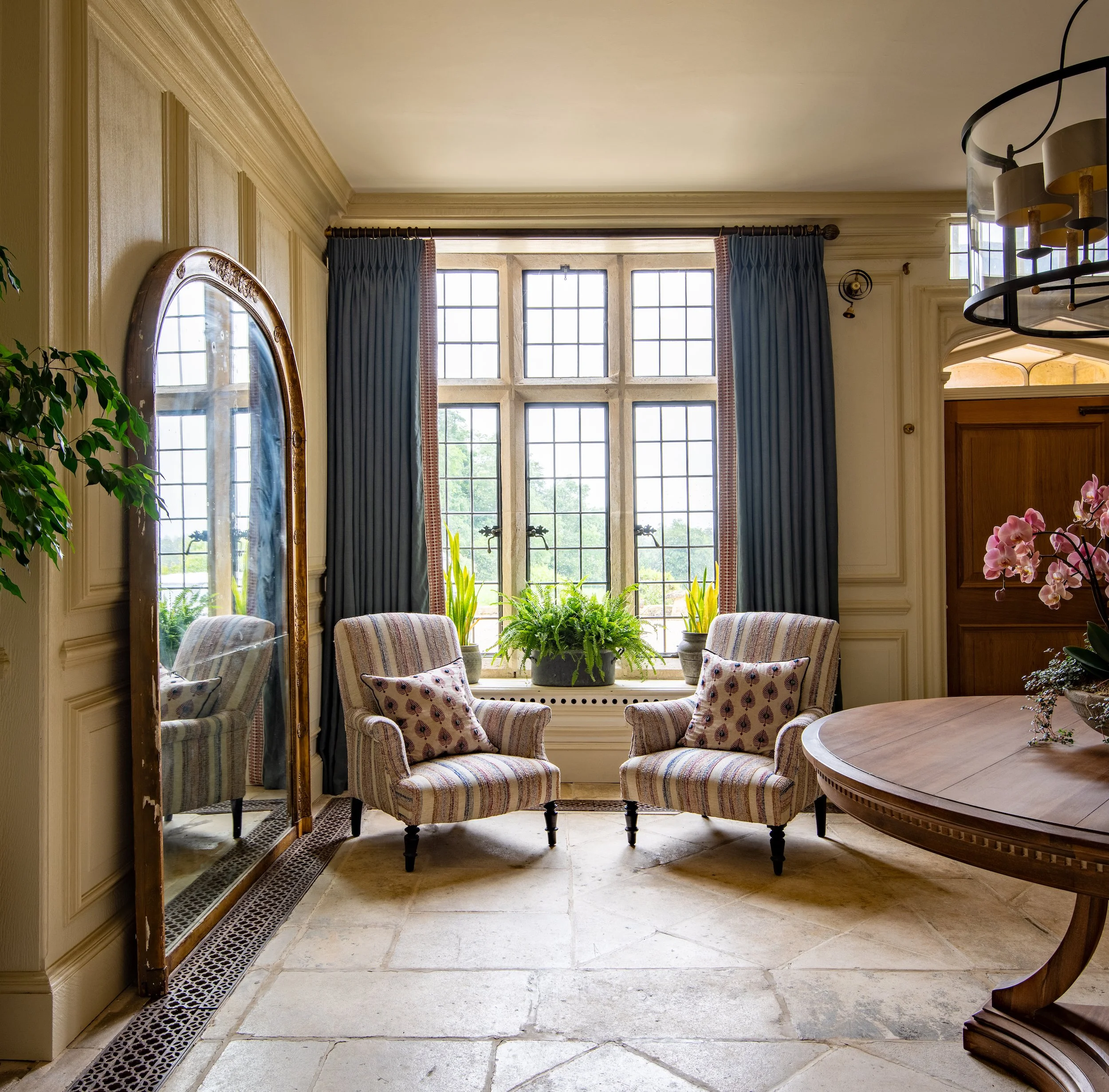 Grand entrance hall designed by MIDA Interiors for a large Cotswolds country estate, featuring original wall panelling, antique floor standing mirror, original stone flooring, antique furnishings and contemporary statement lighting.