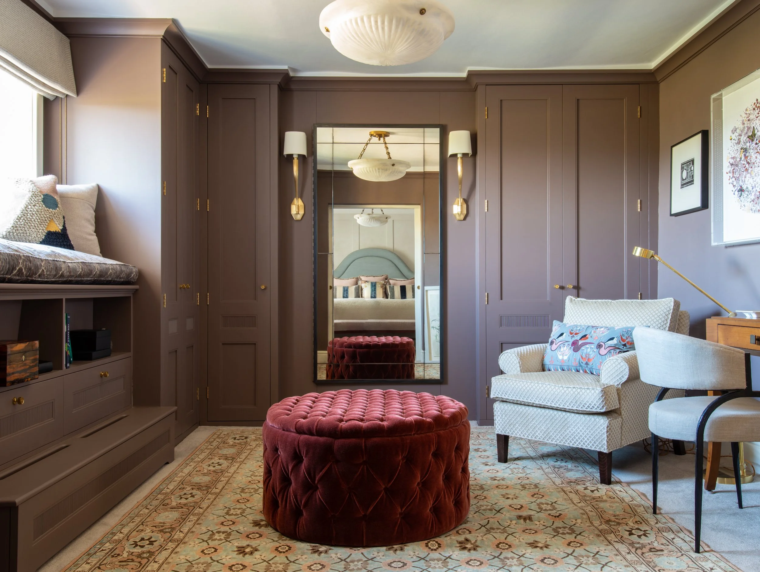 Bespoke dressing room and bedroom interior designed by MIDA Interiors for a Cotswolds country house, featuring fitted cabinetry, hidden storage to rear of large dressing mirror, a window seat, layered lighting, rich colour and custom upholstered furn