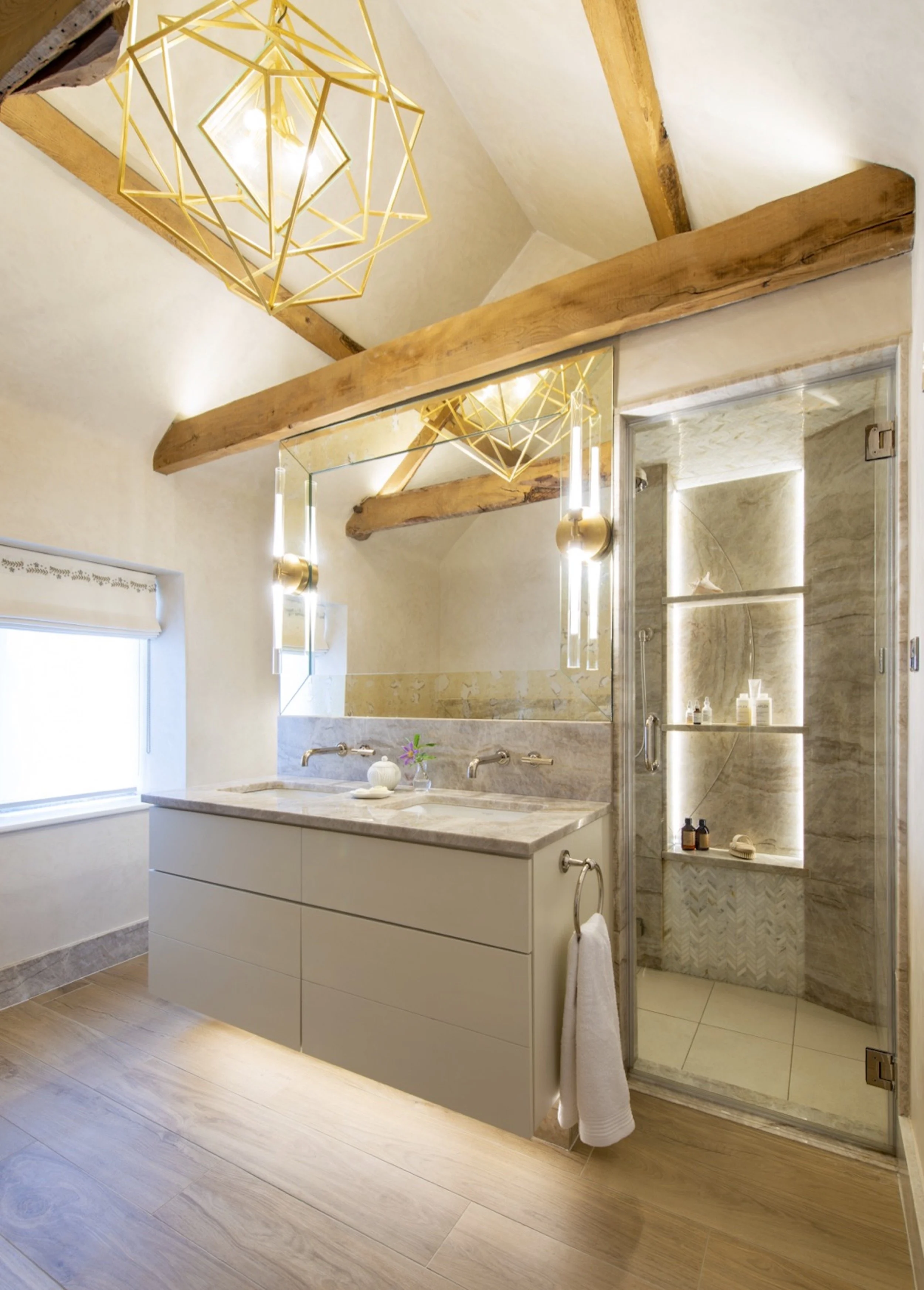 An architectural view of the Cotswold farmhouse principal bathroom, showing the relationship between the wall-hung double vanity and adjoining steam shower. Veined stone surfaces, wall-mounted fittings and vertical brass-and-glass lights frame the ex