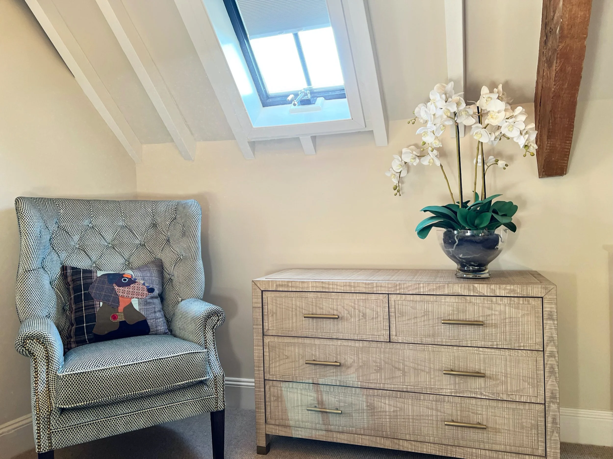 A tailored wingback chair and finely textured chest of drawers create a composed corner within the principal guest bedroom. The playful appliqué cushion, understated brass handles and white orchids bring individuality to the scheme, while the conserv
