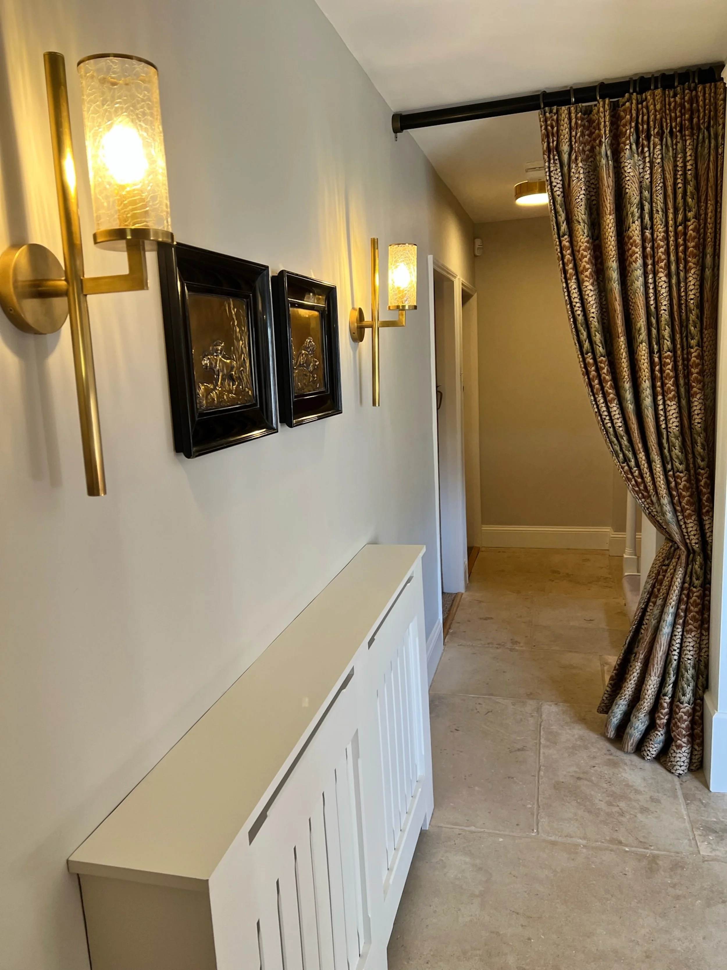 The entrance hall is articulated by contemporary brass and textured-glass wall lights, selected artwork and a full-height patterned curtain that provides a soft division between adjoining spaces. Bespoke fitted cabinetry introduces discreet storage, 