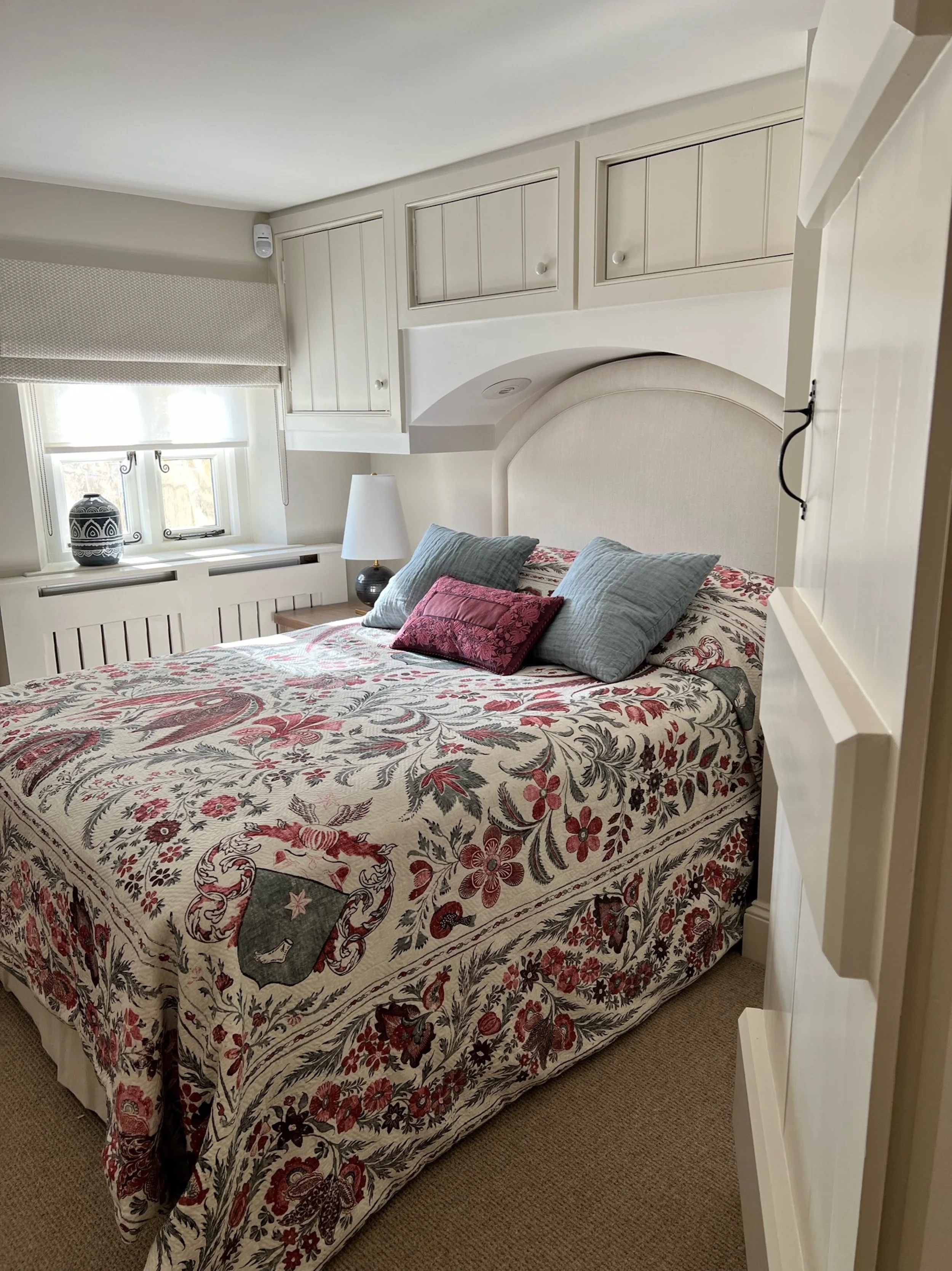This intimate double guest bedroom combines a bespoke upholstered headboard with fitted storage designed around the room’s irregular architecture. A richly patterned bedcover introduces colour and individuality, balanced by muted blue cushions, pale 
