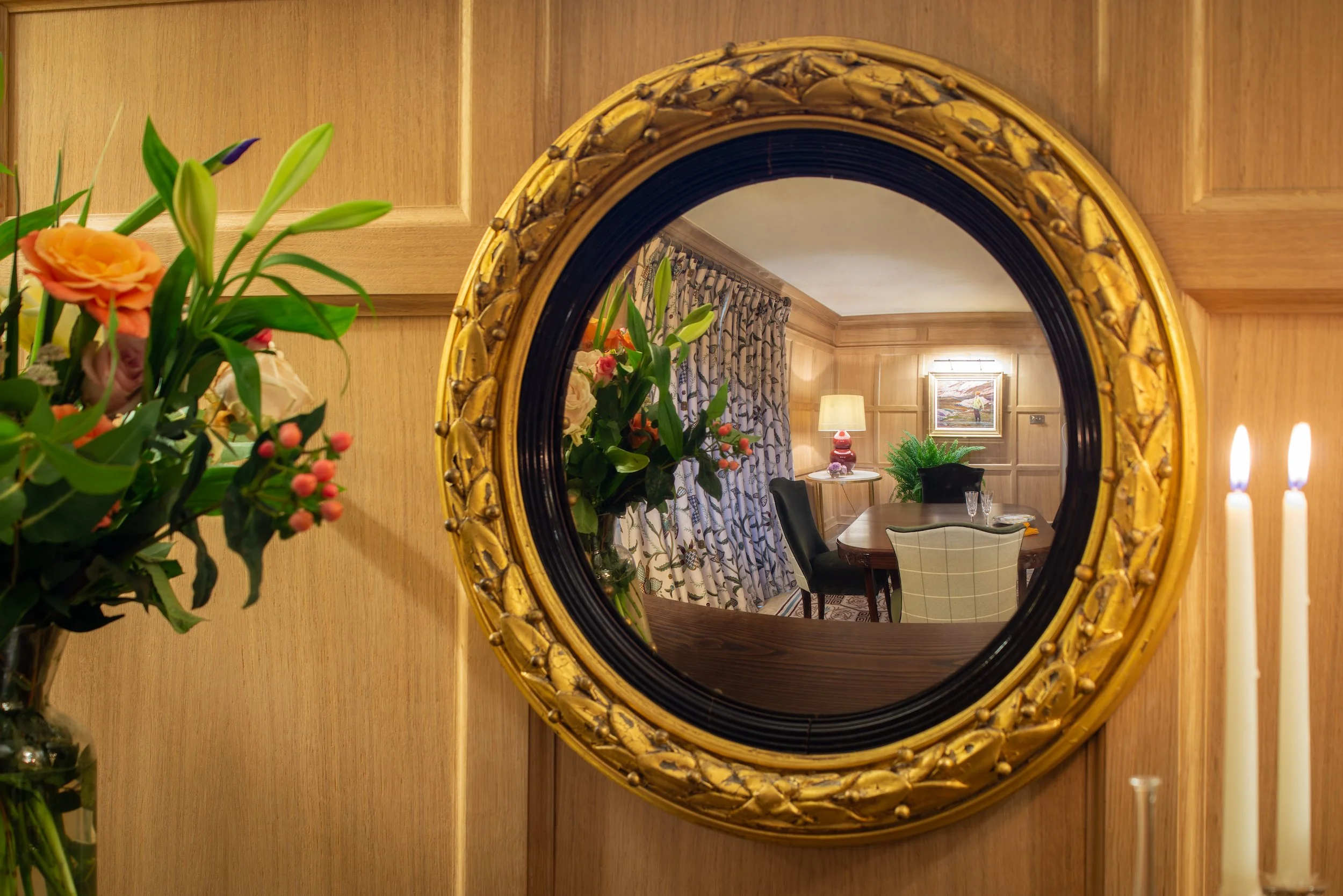A gilt convex mirror mounted against the handcrafted oak panelling reflects the completed Cotswold farmhouse dining room. The view captures the timber dining table, tailored upholstered chairs, crewelwork curtains and layered lighting, illustrating h