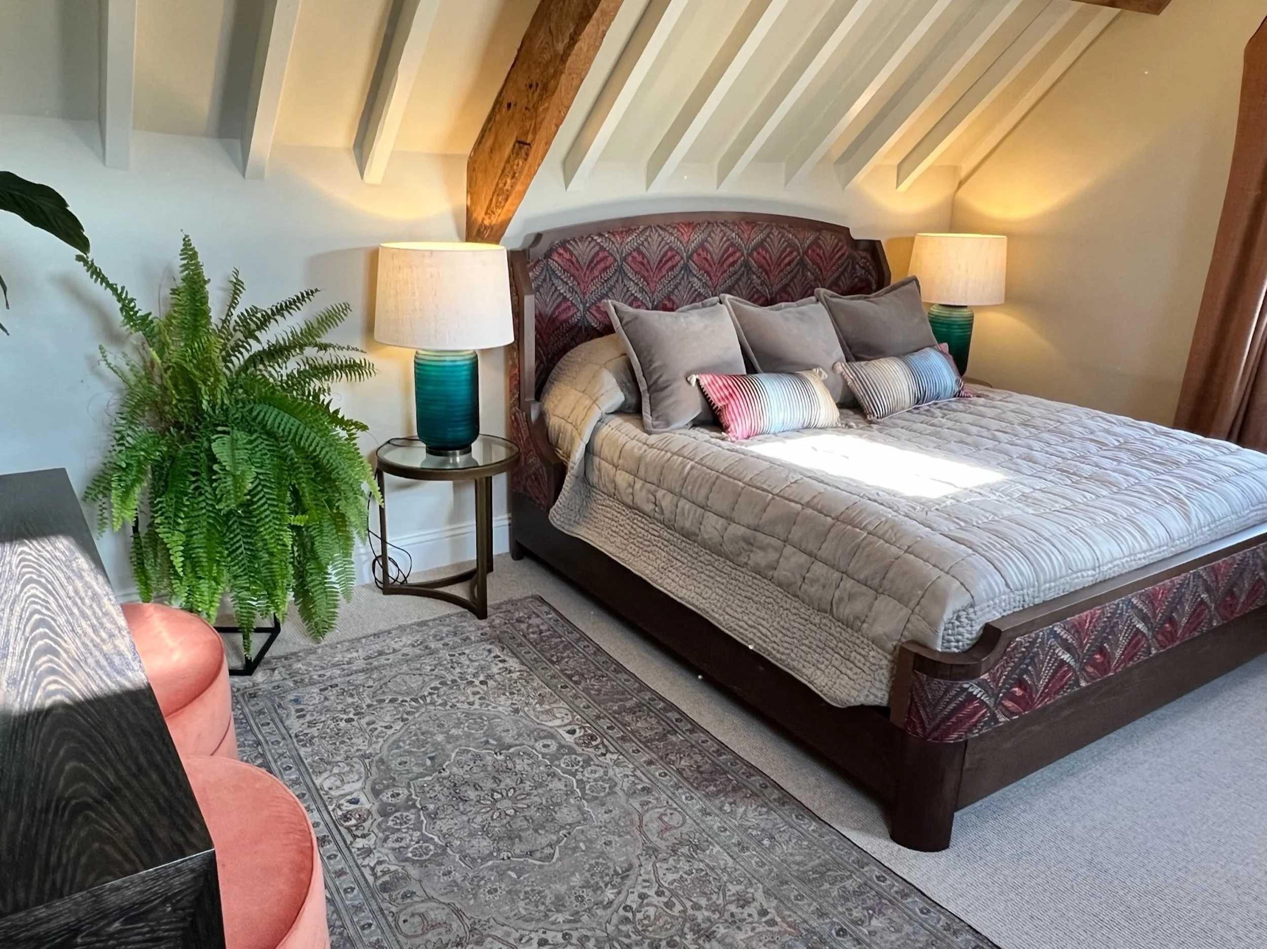 Cotswold country house guest bedroom with an upholstered bed, exposed timber beams, teal lamps and a traditional patterned rug.