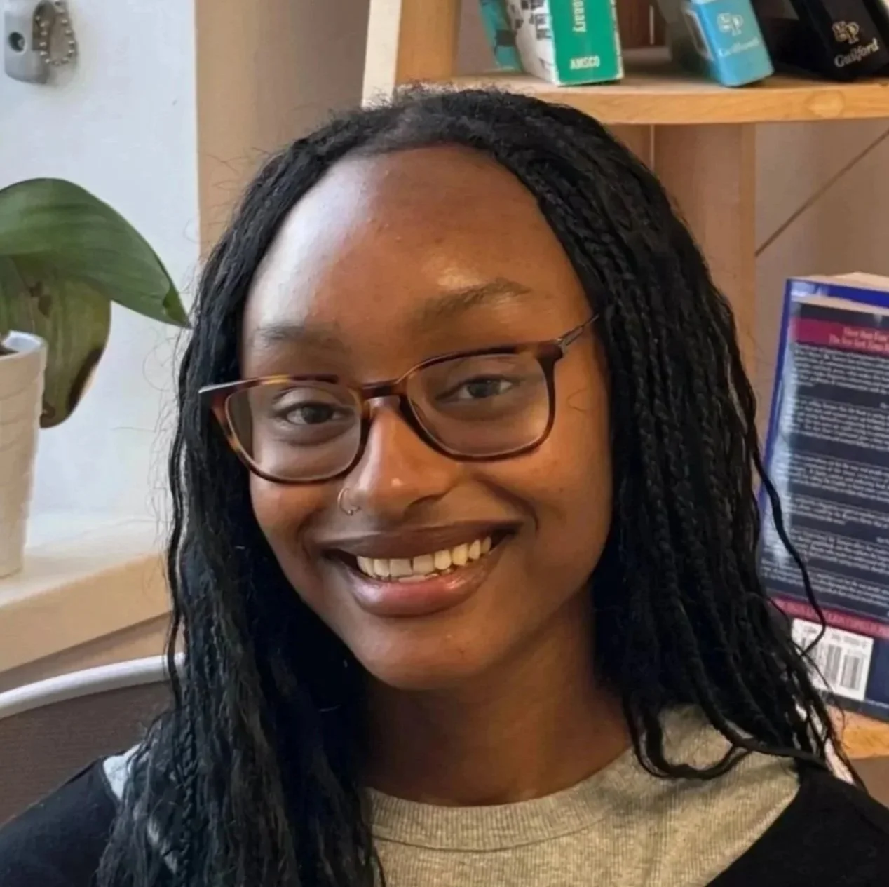 A young woman with dark braided hair, glasses, and a nose ring smiling in an indoor setting with bookshelves and a plant in the background.