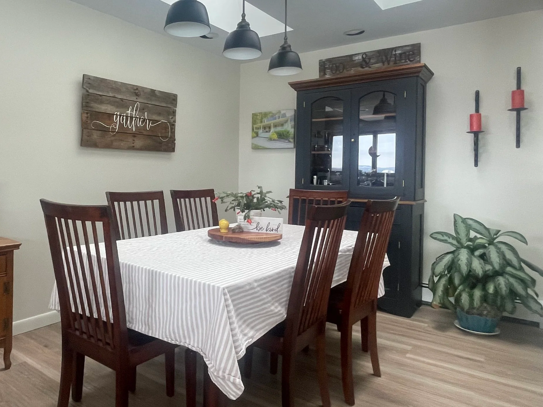 A dining room with a wooden table covered with a white striped tablecloth, surrounded by six wooden chairs, a black hutch with glass doors, a potted plant on the table, and wall decorations including a