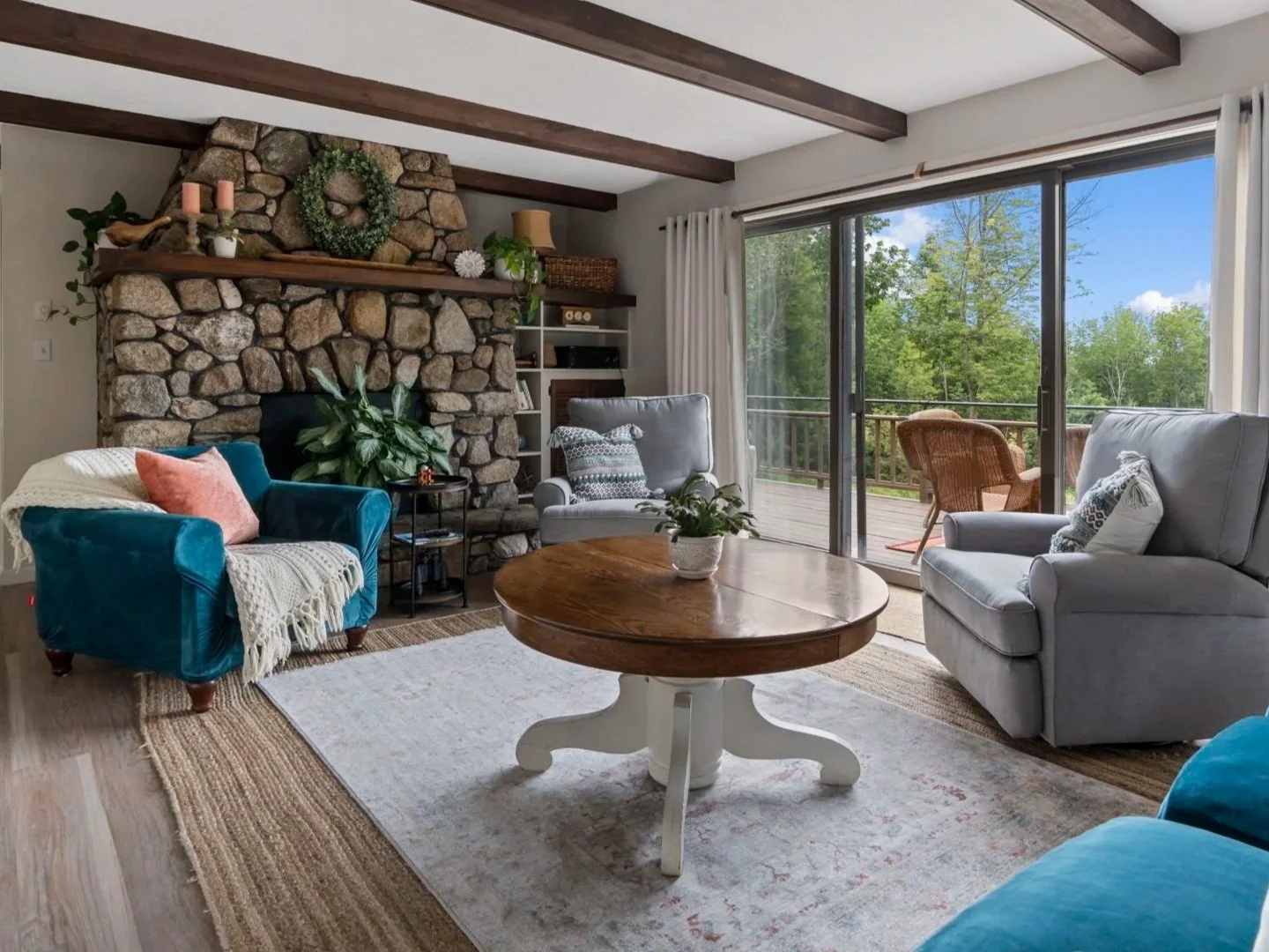 Living room with stone fireplace, blue and gray armchairs, and a sliding glass door leading to a balcony with outdoor seating and green trees outside.