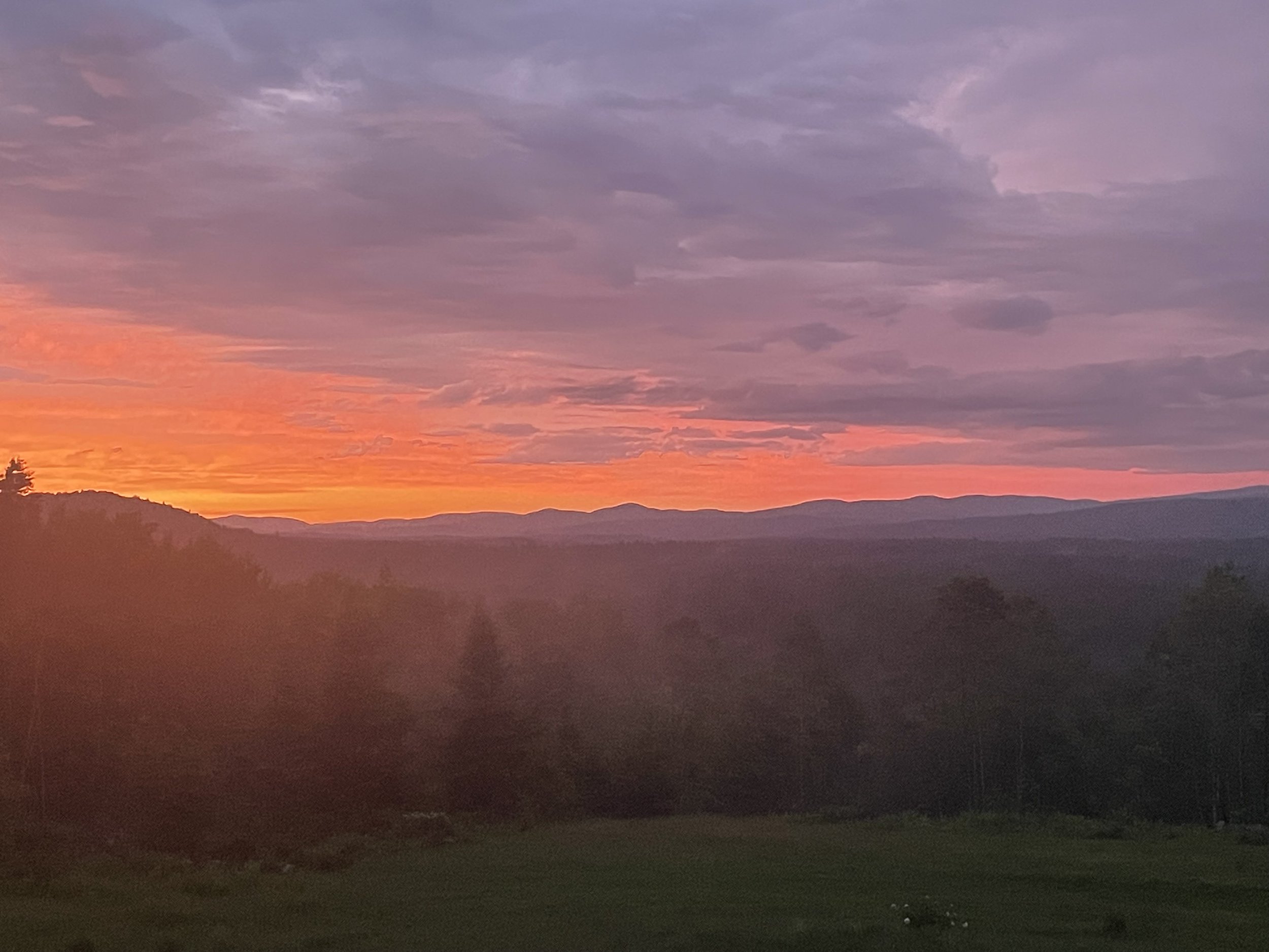 A vibrant sunset over rolling hills and trees, with clouds in the sky painted in shades of purple, pink, and orange.