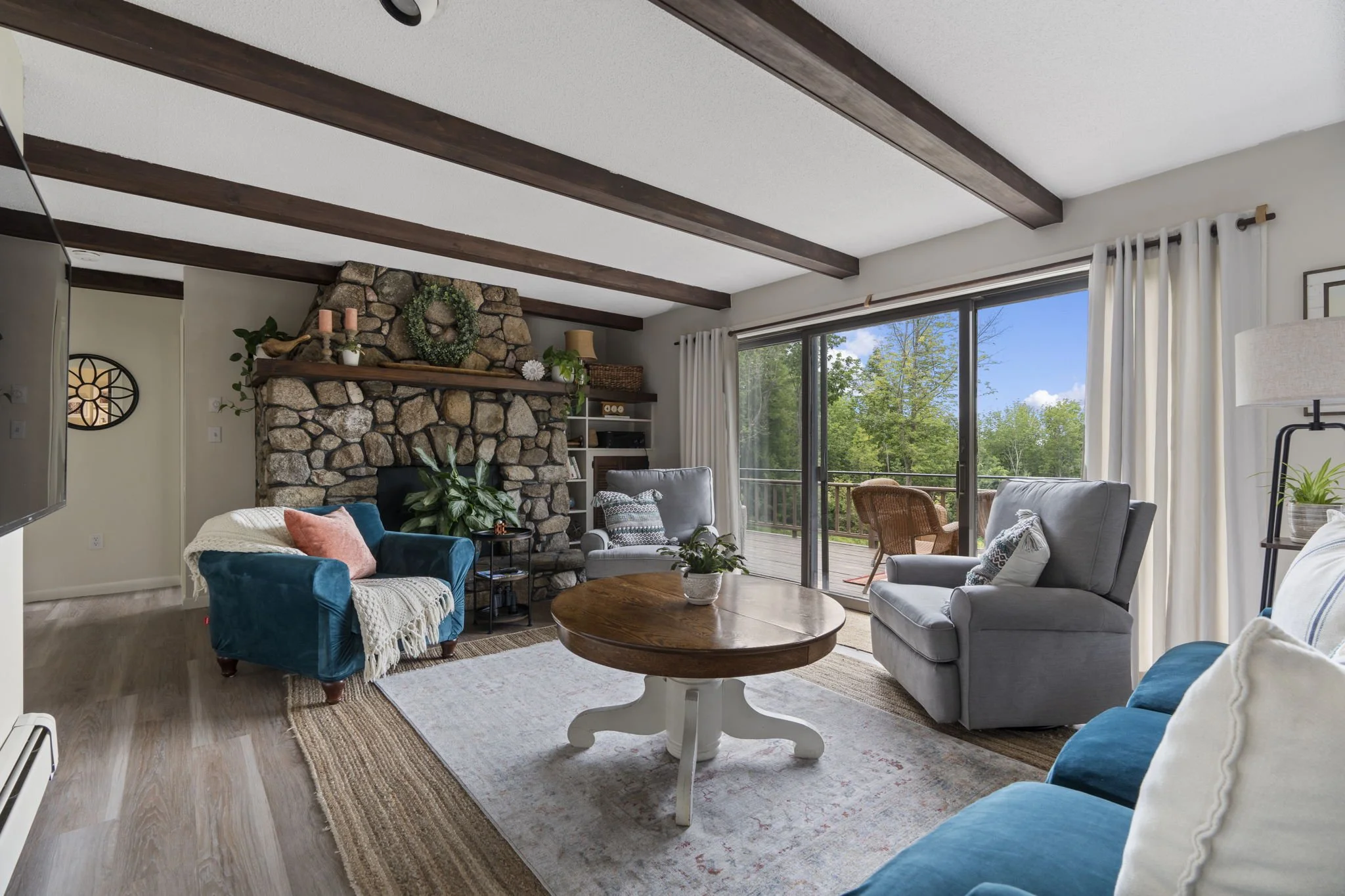 Living room with a stone fireplace, blue and gray armchairs, a round wooden coffee table, and a large sliding glass door leading to a deck with outdoor seating. Decor includes plants, pillows, and curtains, with trees visible outside.