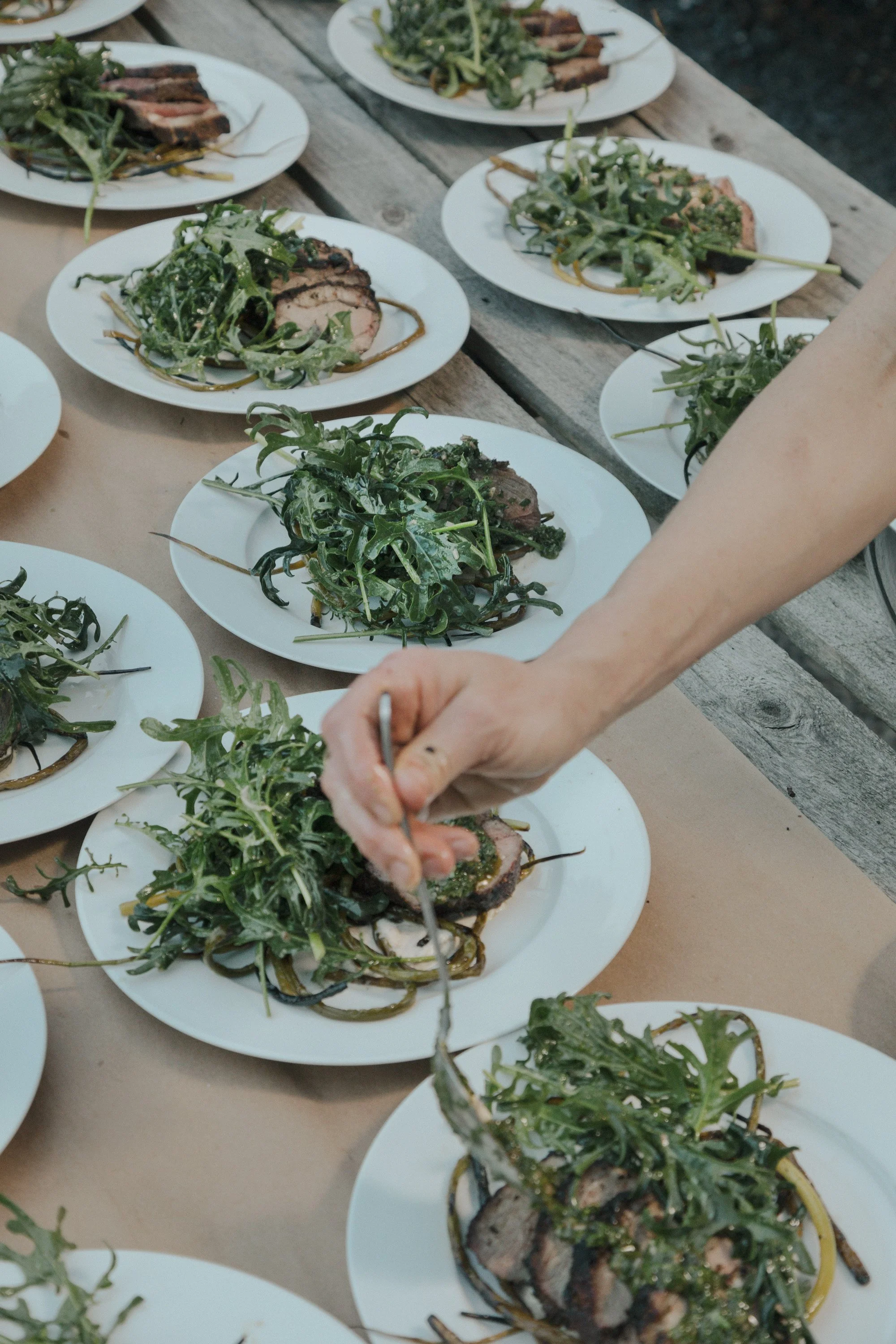 A wooden table is covered with white dishes filled with grilled lamb and greens. A hand in the bottom half is dressing one of the plates with sauce.