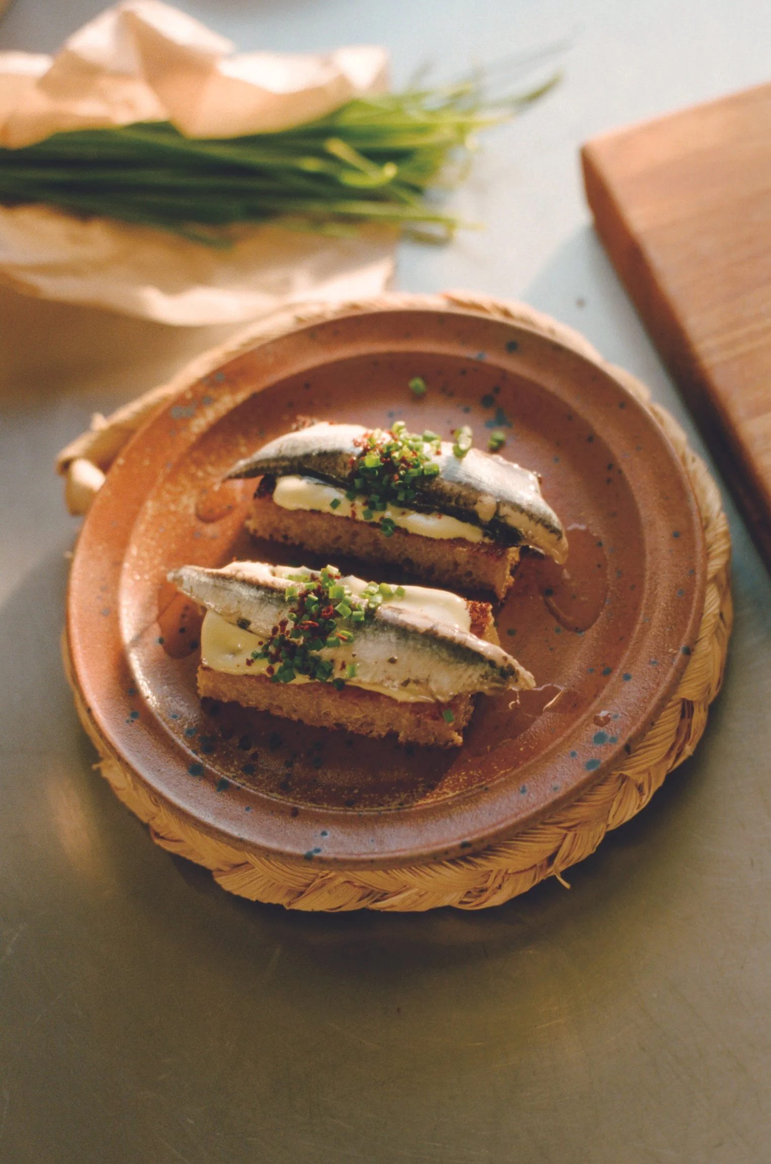 Two small boquerones toasts on a hand made rich brown ceramic plate. Sun is streaming in and the anchovies are slick and topped with chives and chili flakes.