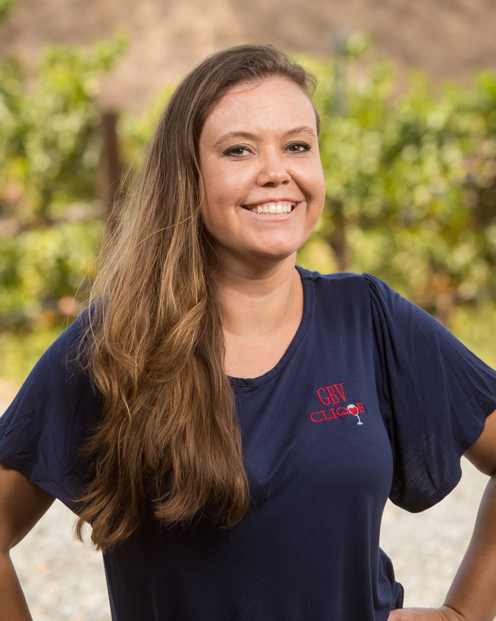 Smiling woman wearing a black T-shirt with "GBV Clique" logo, standing outdoors with trees in the background.