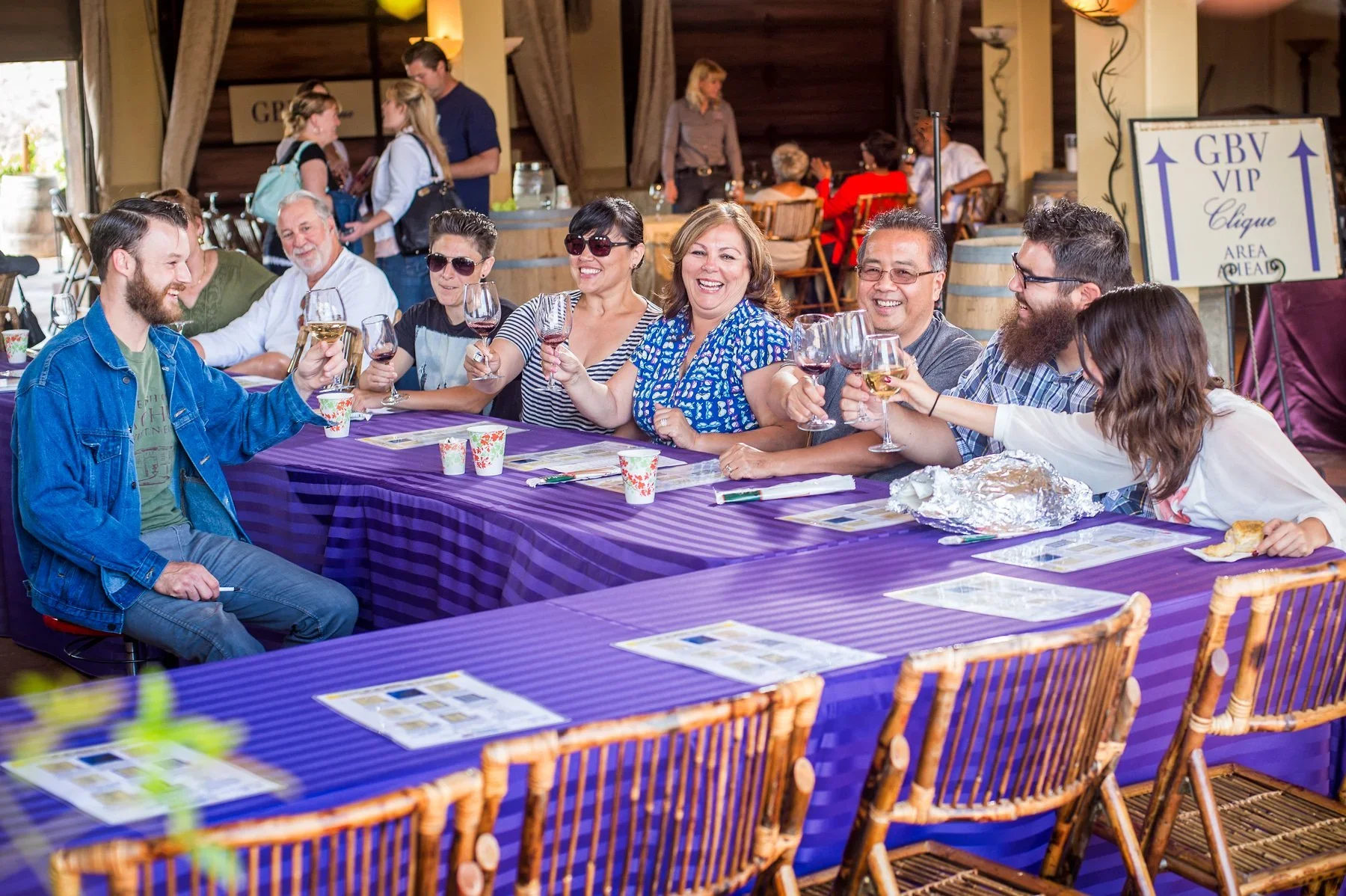 Group of people at a table toasting with wine glasses, seated in a winery setting with purple tablecloths.