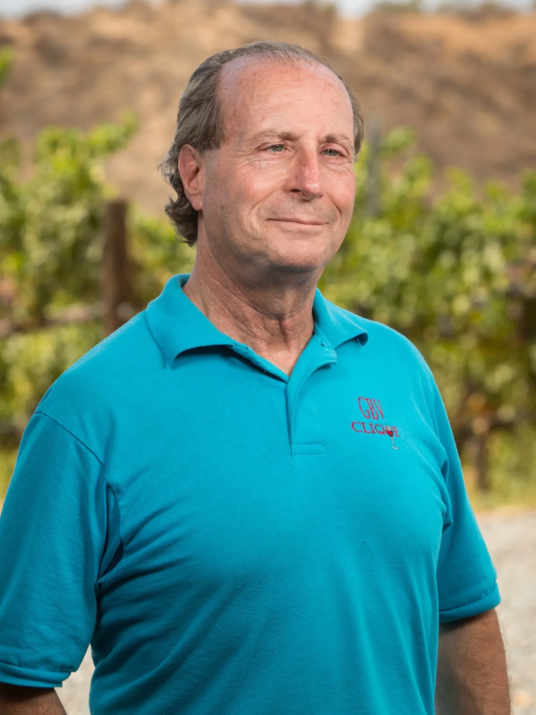 Man wearing a blue polo shirt standing in an outdoor setting with blurred greenery in the background.