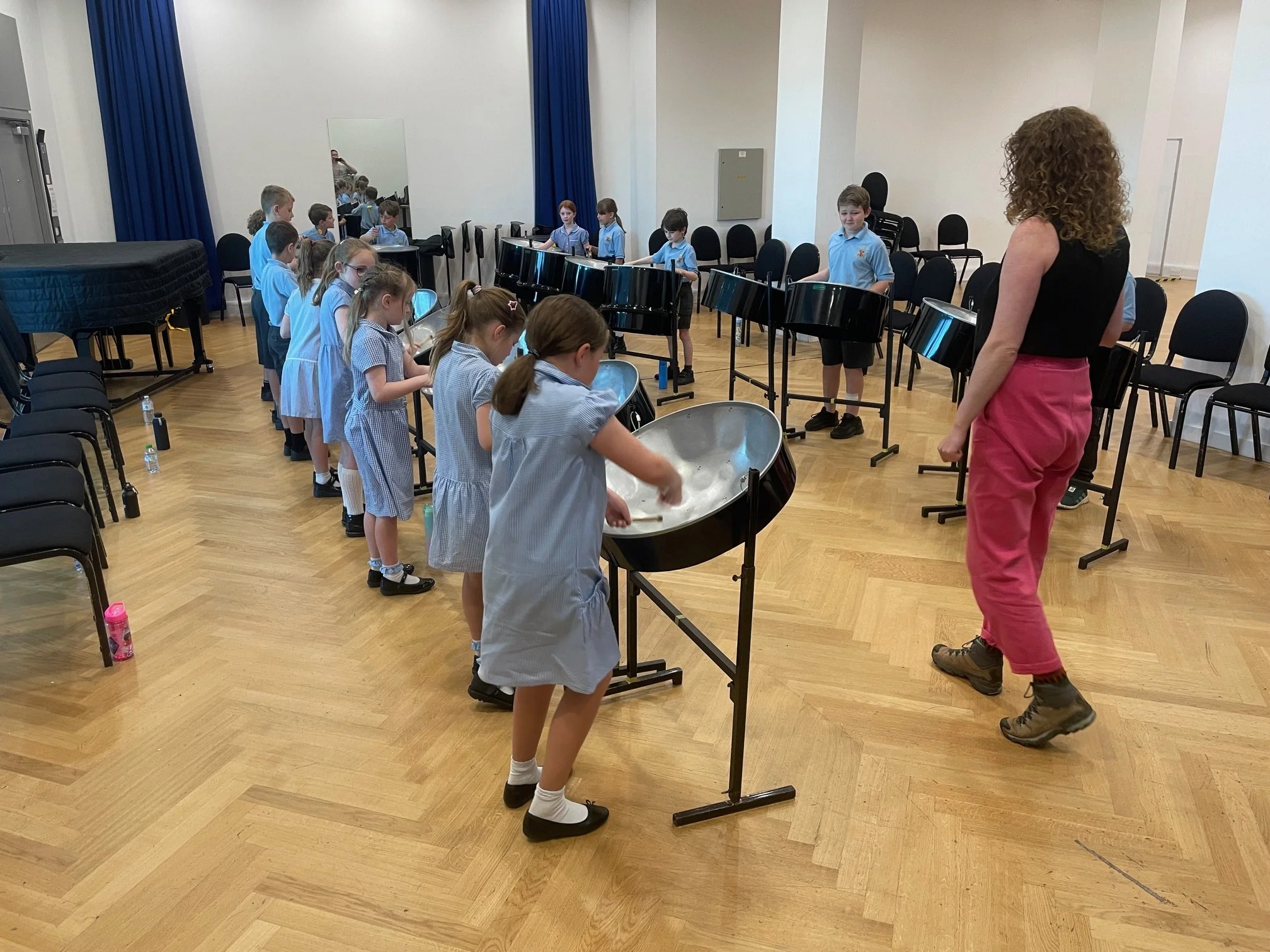 Two children in a classroom playing blue ukuleles, sitting at a desk with other students in the background, and school supplies on the desk.