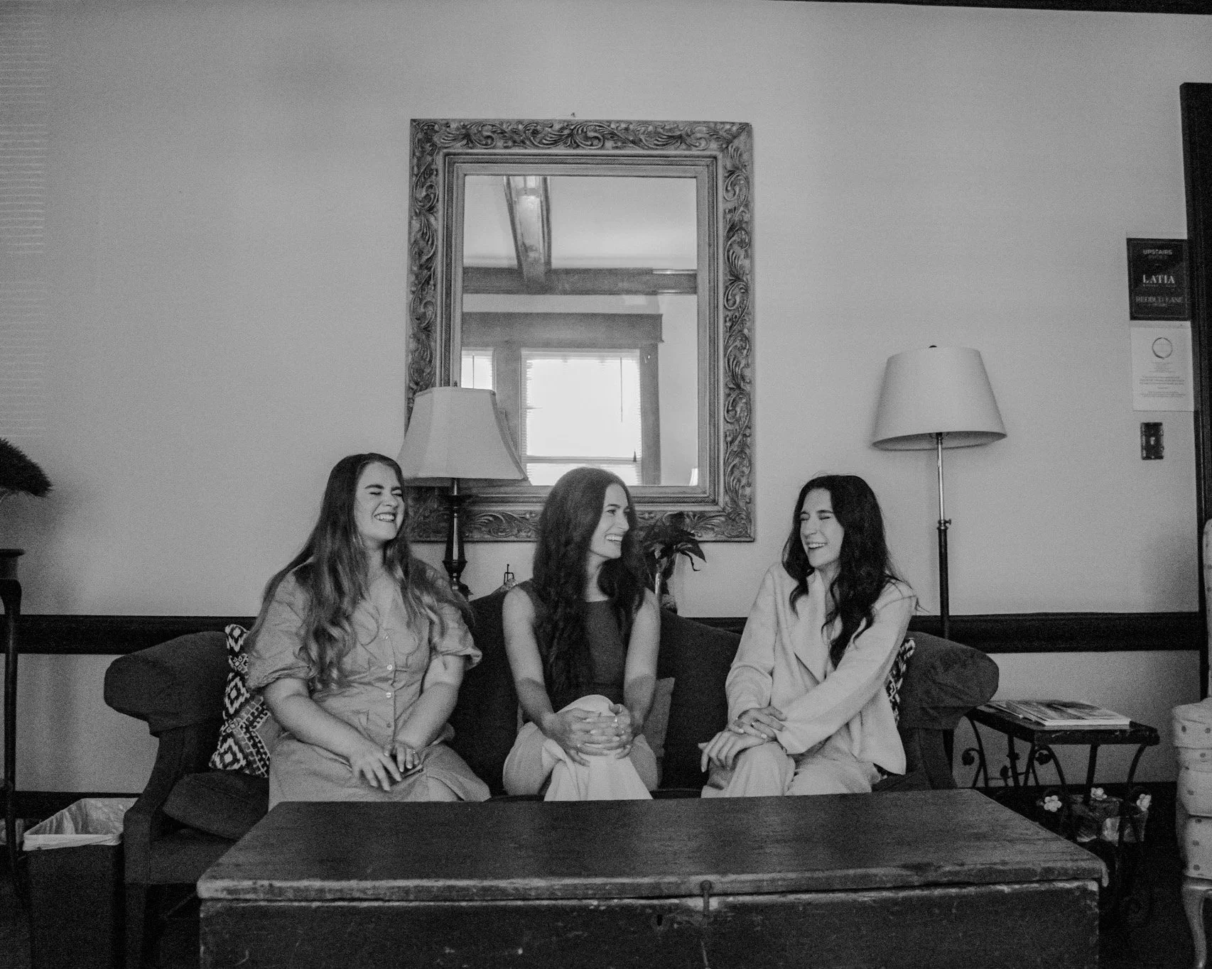 Three women sitting on a couch in a living room, smiling and having a conversation. A mirror hangs on the wall behind them, with a window reflected in it. There are lamps and a side table with magazines.