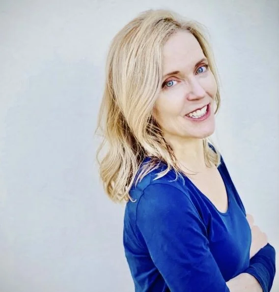 Cate Allen is a light-skinned woman who is in her 40s. She has blonde hair and blue eyes, and is wearing a blue blouse. She has her arms crossed.