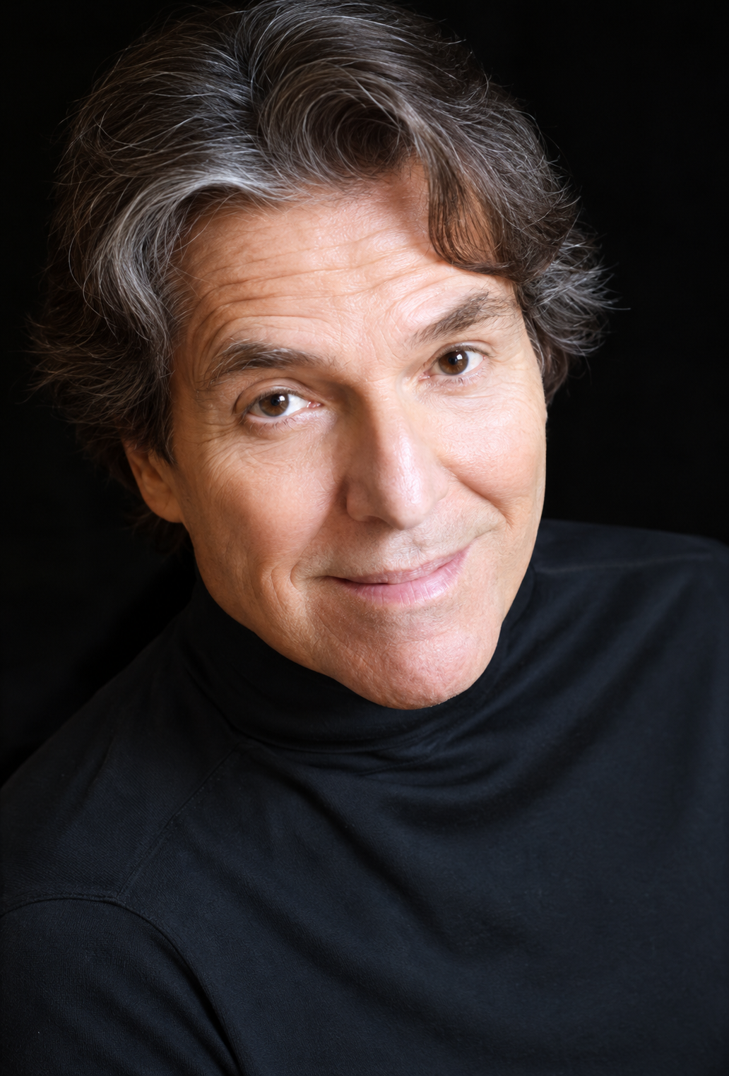 A light-skinned man, 60s, wavy gray hair and dark eyes, he’s wearing a black top.