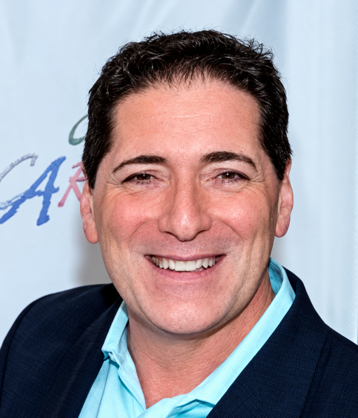 Nicholas Viselli, (Artistic Director). Light-skinned man, 60s, brown eyes and short dark hair. He’s wearing a light blue shirt with a black blazer and he is smiling.