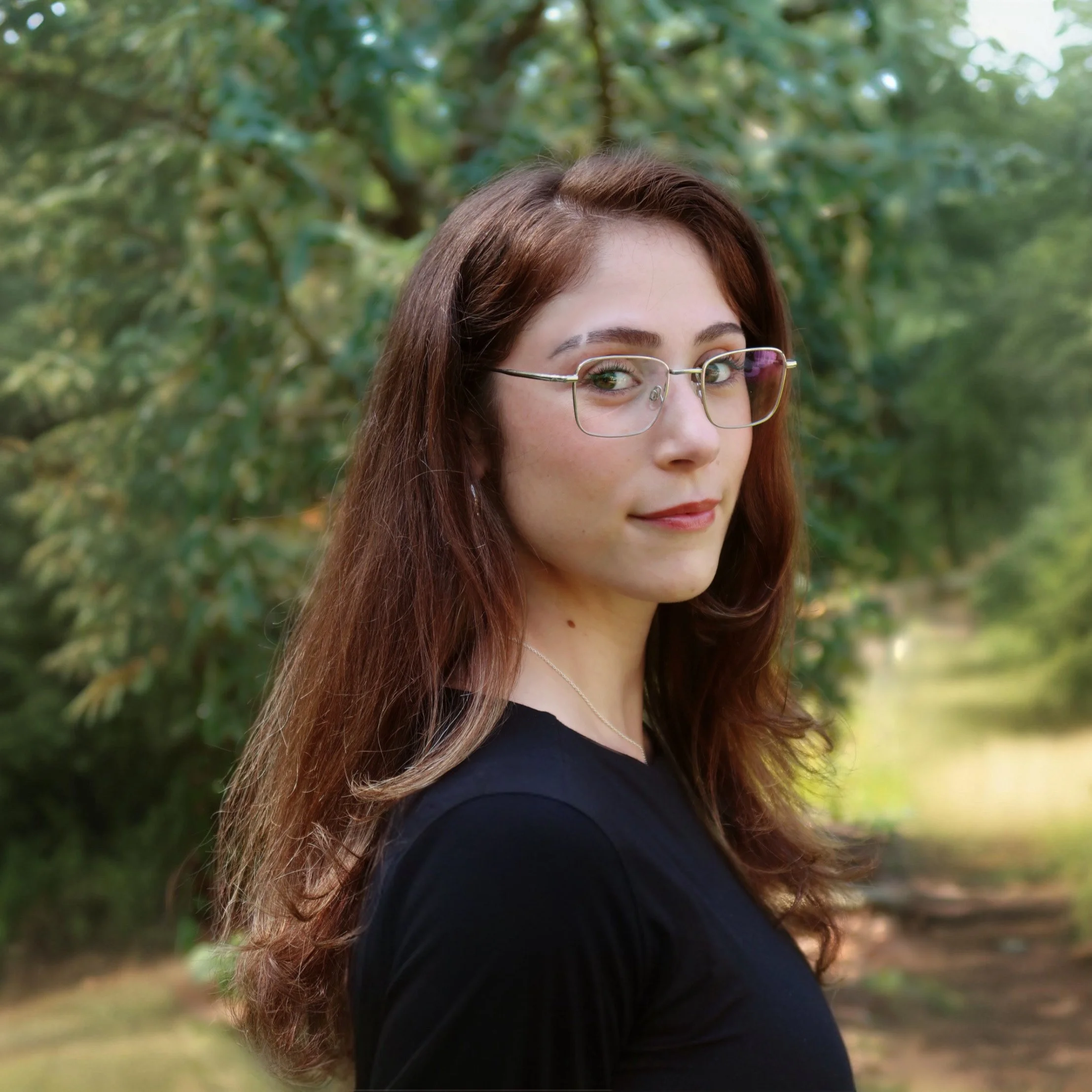 Abrianna Sigel, a light-skinned woman, 20s, long brown hair, glasses, wearing a black shirt, standing outdoors with a large tree behind her.