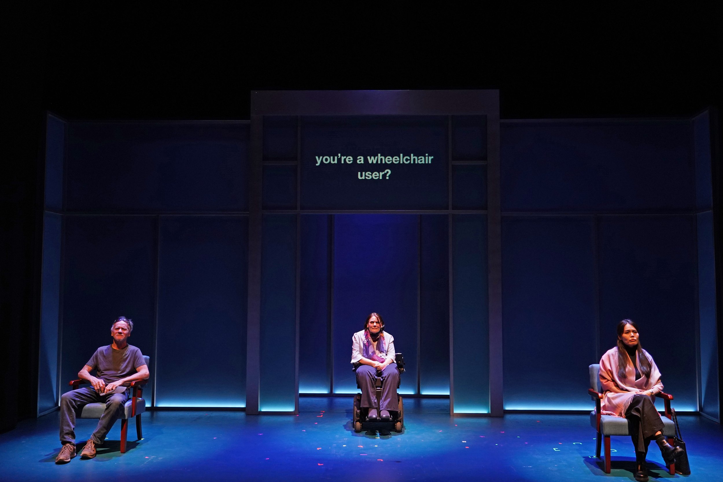 Three light-skinned people sit on a stage under purple-blue lighting; they are all wearing casual clothing, and the person in the center uses a wheelchair. Above them, projected text reads, “You're a wheelchair user?”