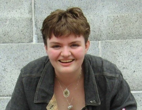 Jo Dedolph is a light skinned nonbinary individual in their 20s with short brown hair. They’re smiling and wearing a dark jacket and layered necklaces, in front of a light gray stone wall.