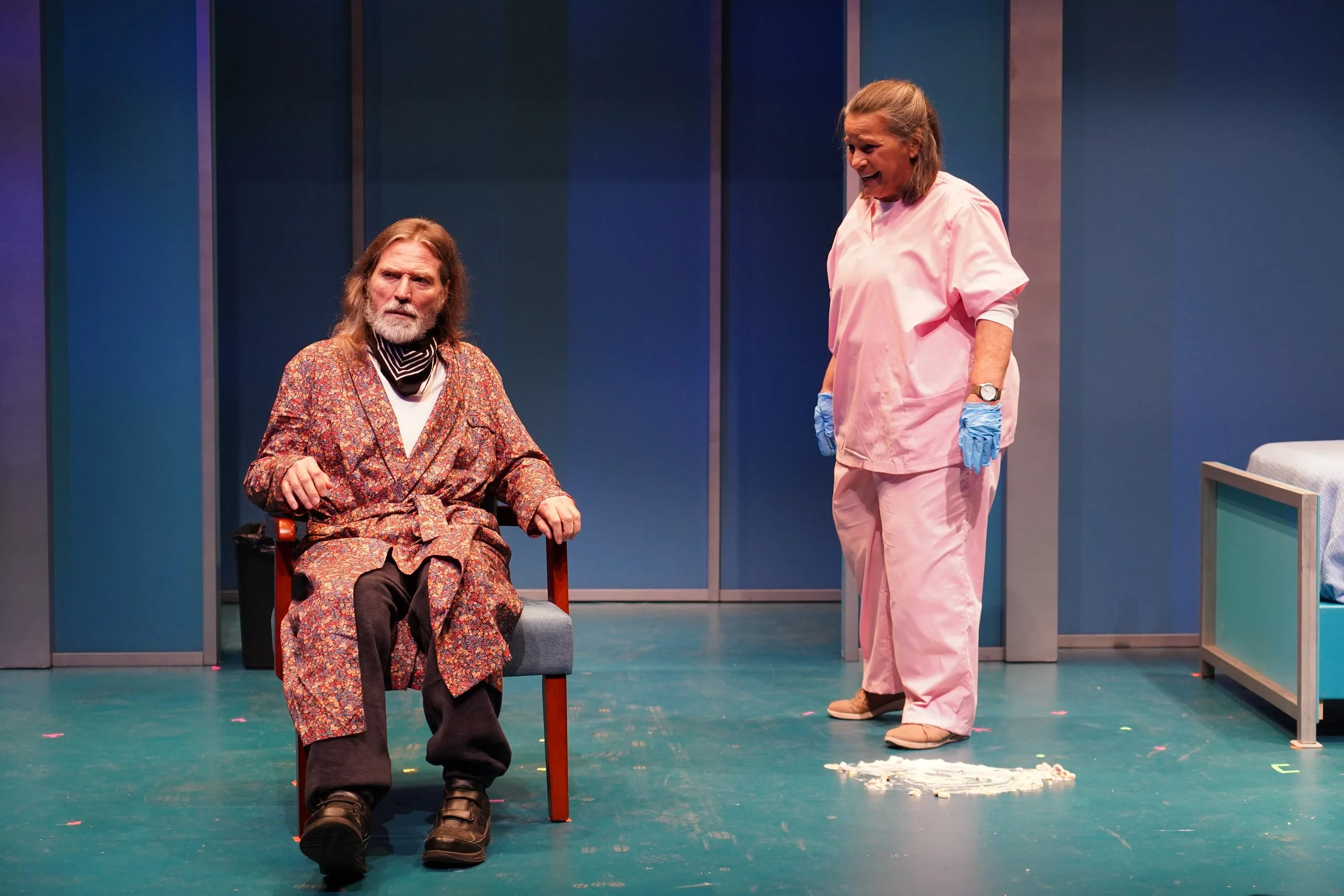 An older, light-skinned man in a patterned robe sits on a chair while a woman in pink scrubs and blue gloves stands nearby, looking at a mess on the floor beside a bed in a brightly lit room.