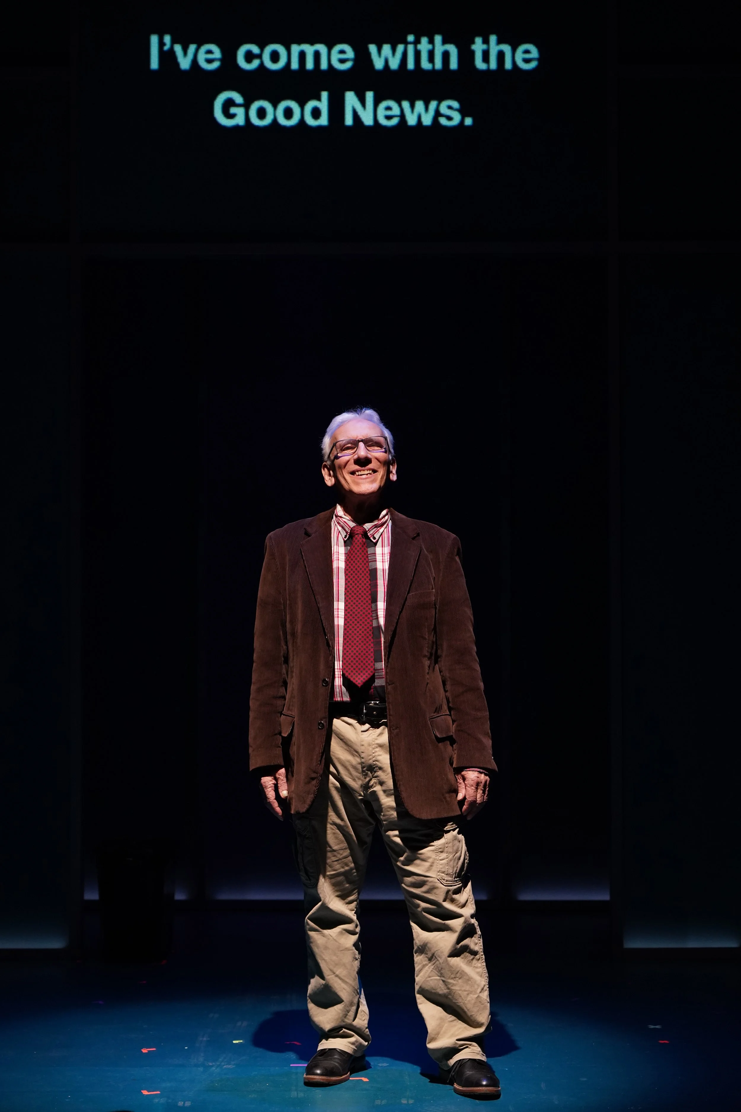 An older light-skinned man in glasses, a red plaid shirt, red tie, brown jacket, and khaki pants, stands smiling on a dimly lit stage. Above him, text reads, “I’ve come with the Good News.”