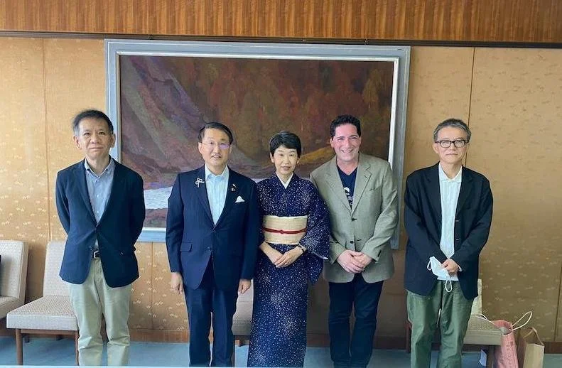 A picture of five people who are standing in line and wearing formal attire. Behind them, there is a painting on a wall.