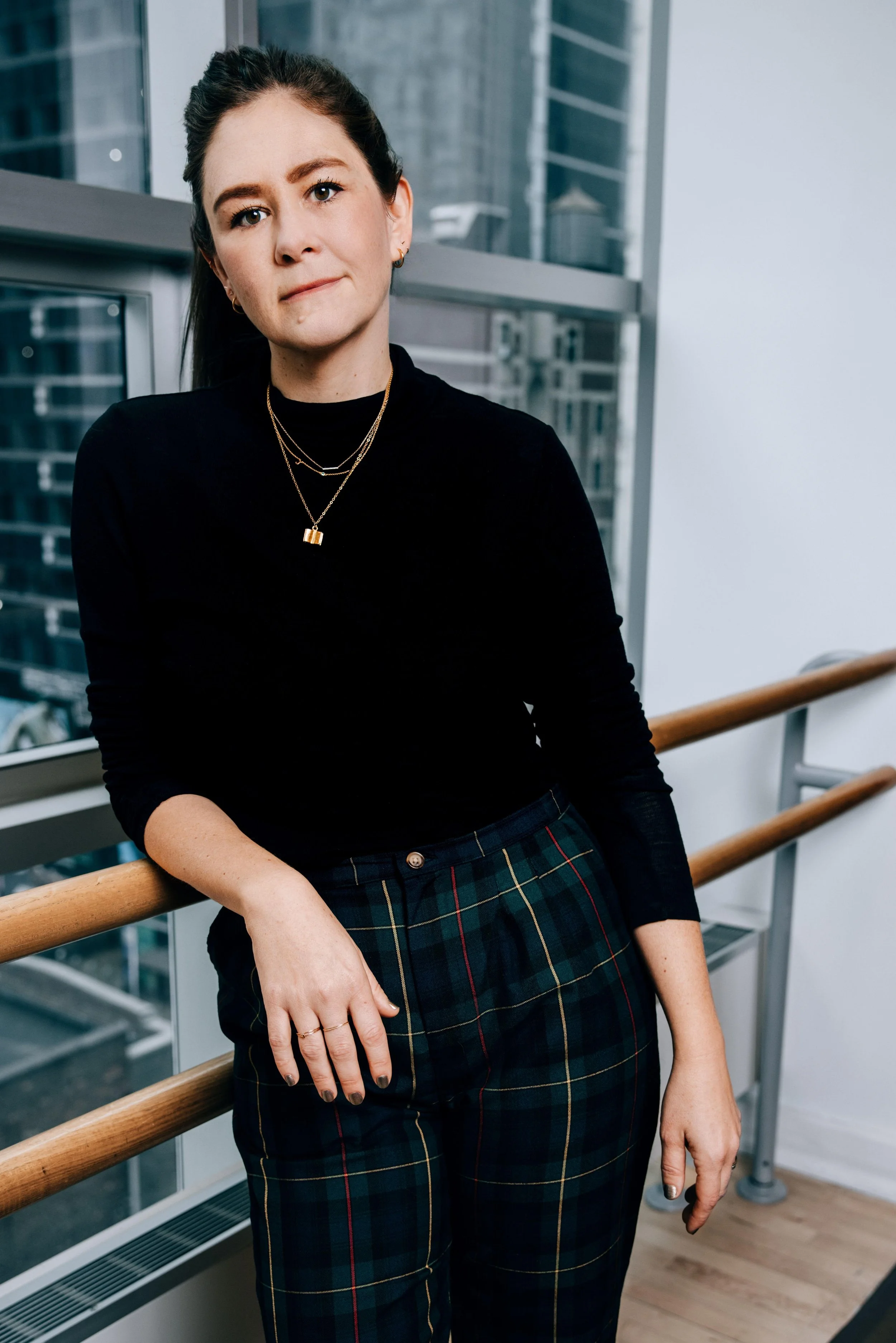Bekah Brunstetter is a light-skinned woman who is in her late-30s. She has dark hair in a ponytail with dark eyes, and is wearing a black shirt with necklaces and plaid pants. She is leaning against ballet bars.
