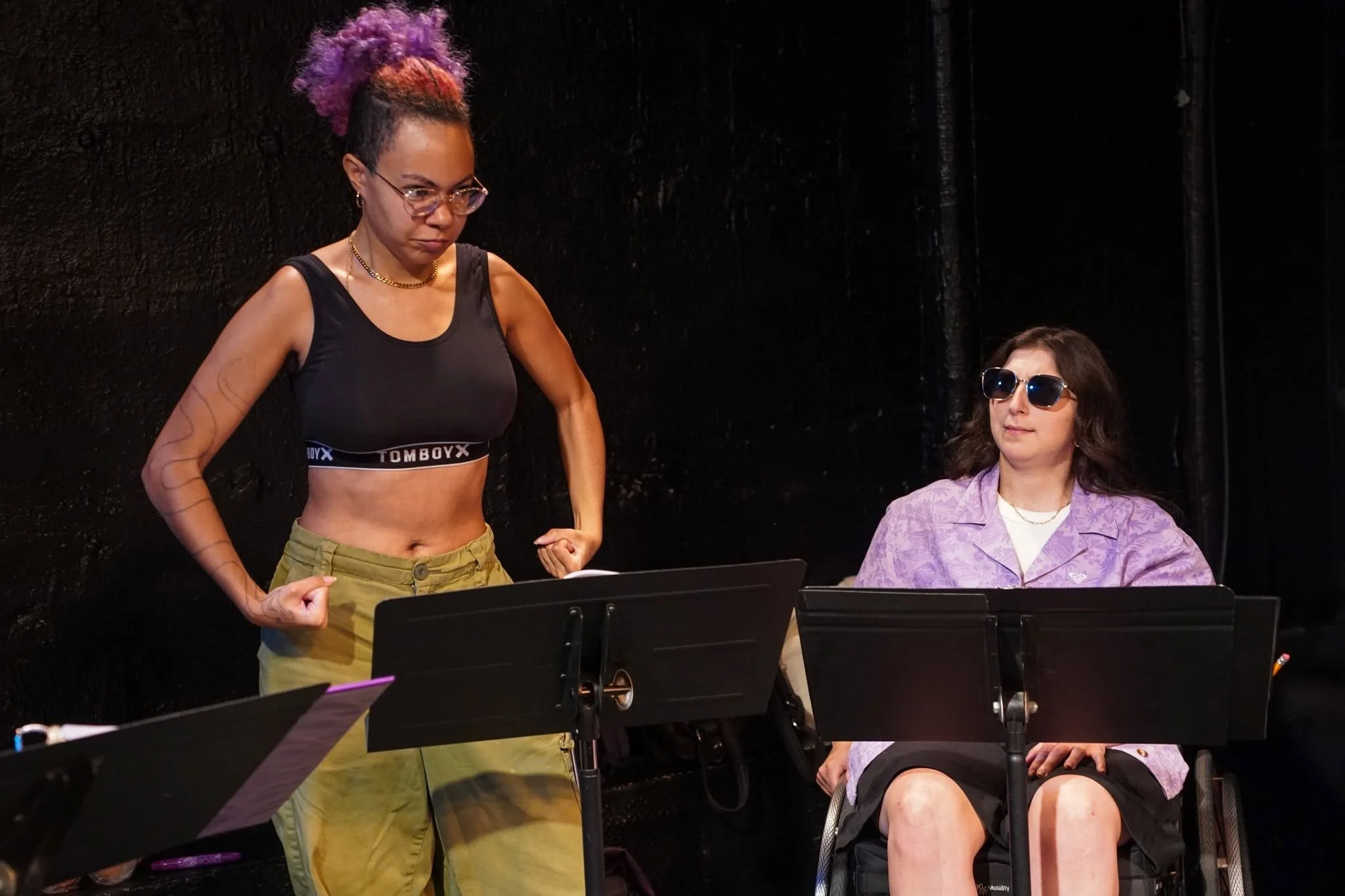 A person with purple hair wearing a black crop top and green pants flexing muscles. Beside them, there’s a seated individual in a wheelchair with a purple shirt.