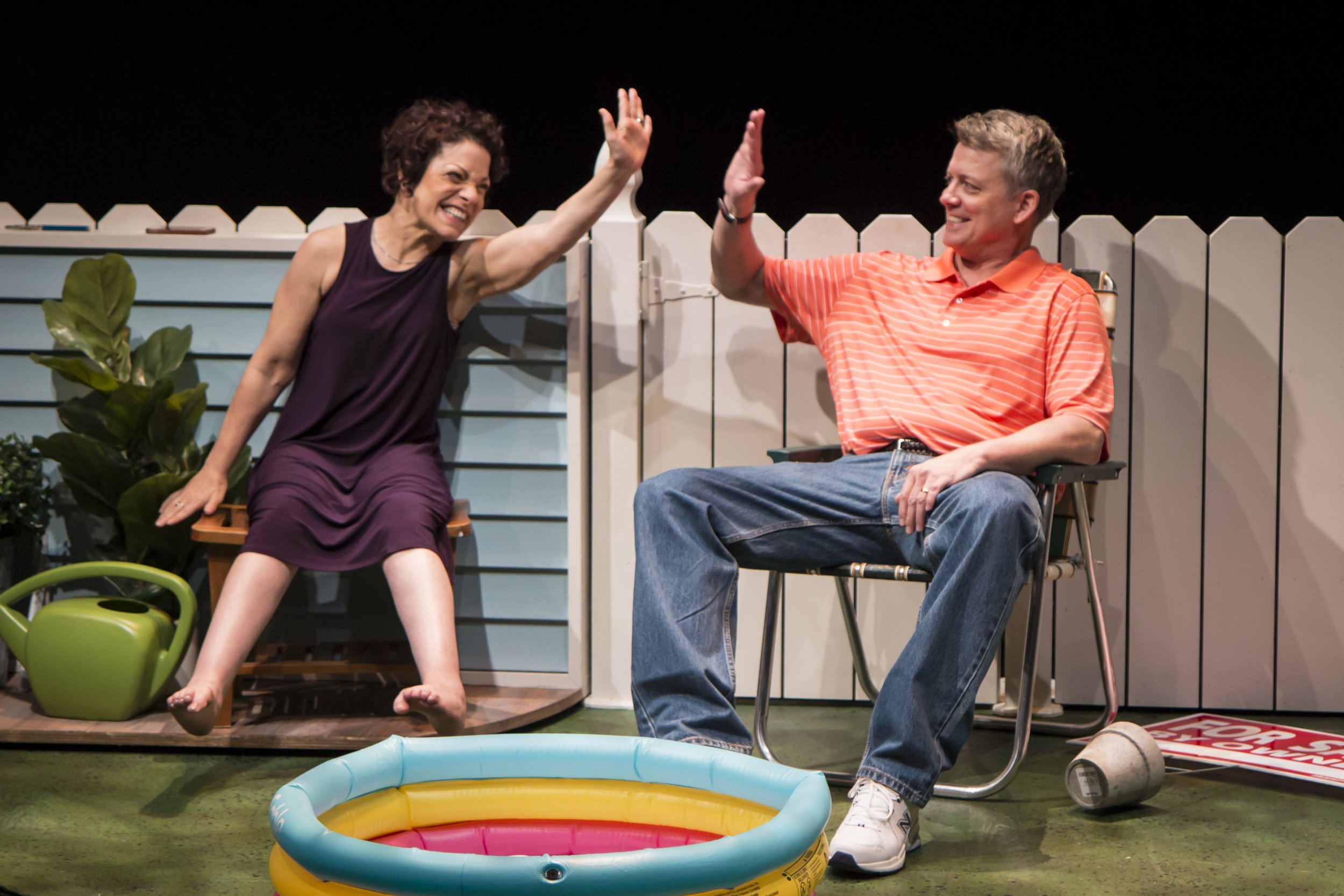 2 actors sitting in a garden in front of a small plastic pool for children. They smile and are about to do a high five. Both are light-skinned. A woman in her 40s, with short brown hair. A man in his 50s with short grey hair.