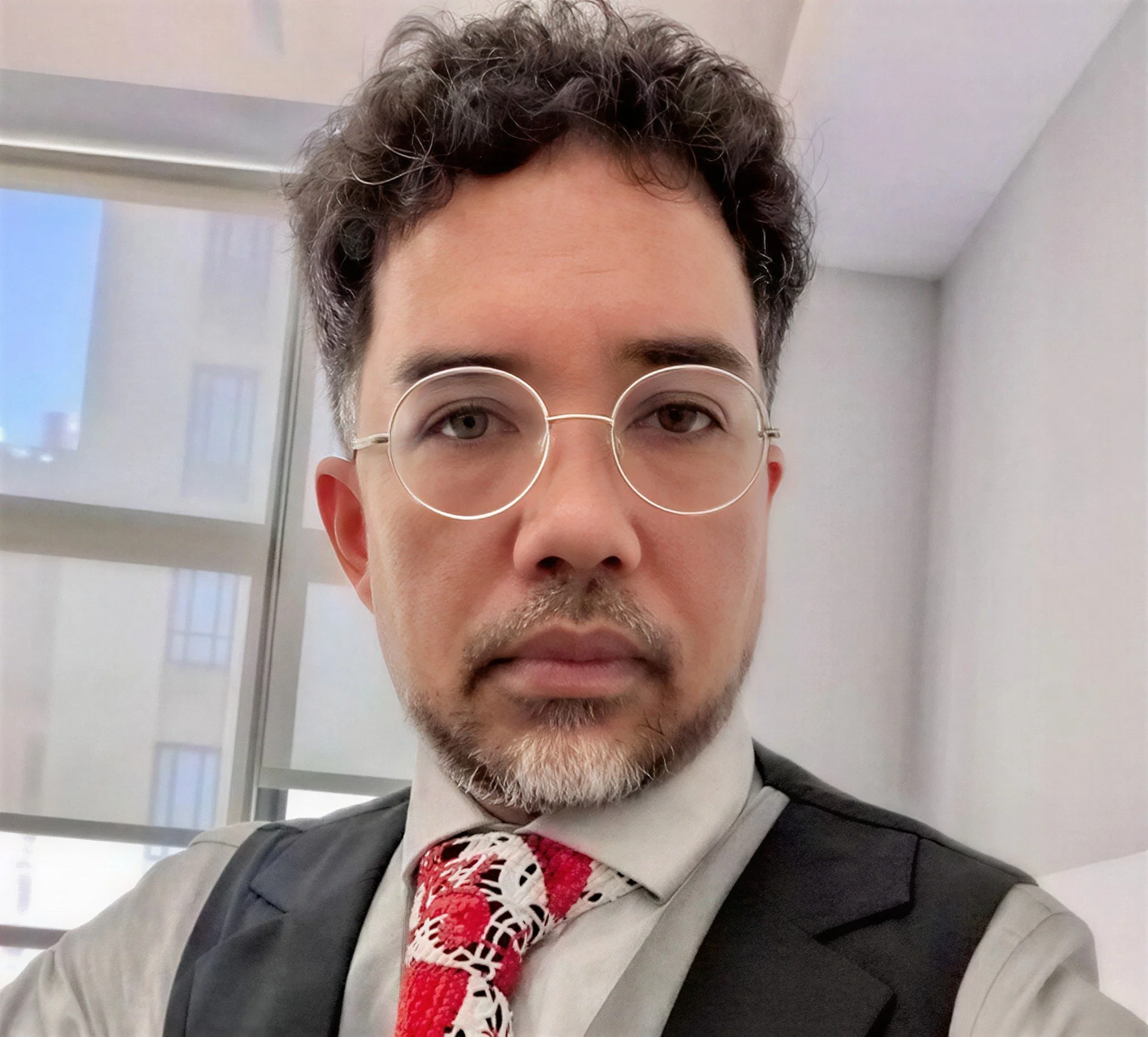 A light-skinned man with hazel eyes, 50s, has curly dark hair, glasses, and a goatee, wears a gray shirt, black vest, and a red patterned tie.