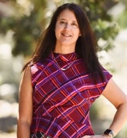 Woman with long dark hair wearing a purple and red plaid sleeveless top standing outdoors with trees in the background.