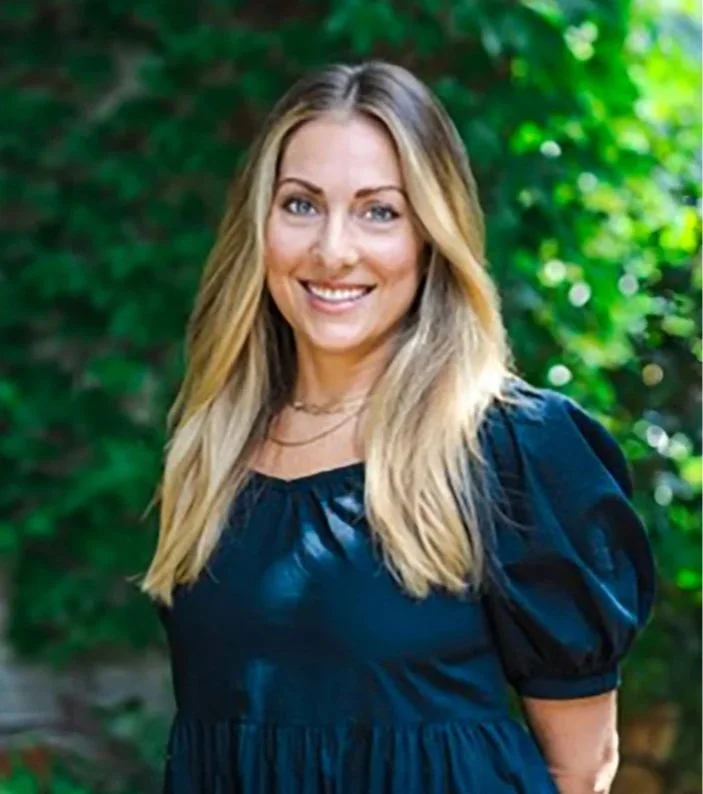 Smiling woman with long blonde hair standing outdoors with green foliage background.