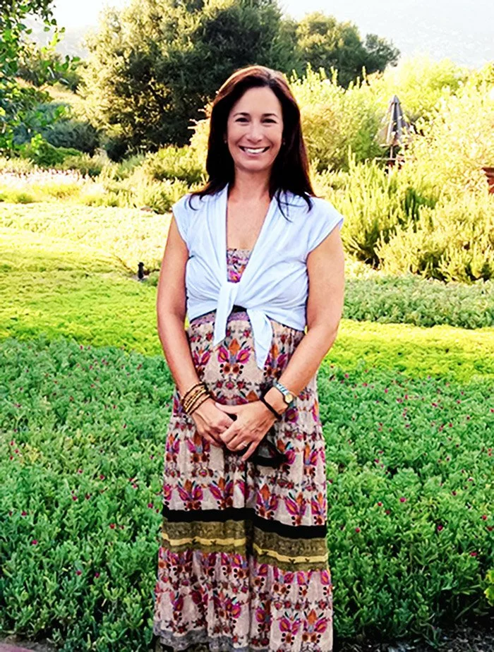 A woman with long dark hair smiling outdoors in a garden with green plants and trees, wearing a patterned dress and a light blue top tied at the front.
