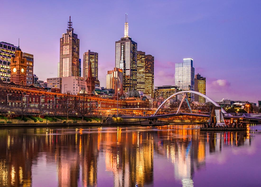 City skyline at dusk with tall skyscrapers, a bridge over a river, and reflections on the water