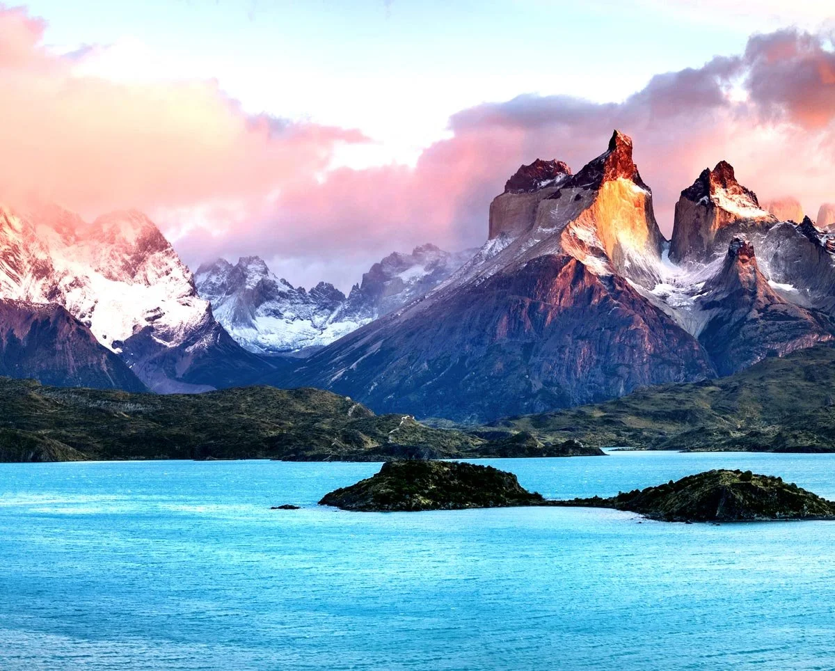 Scenic landscape of snow-capped mountains with a colorful sky, and a bright blue lake with small islands in the foreground.